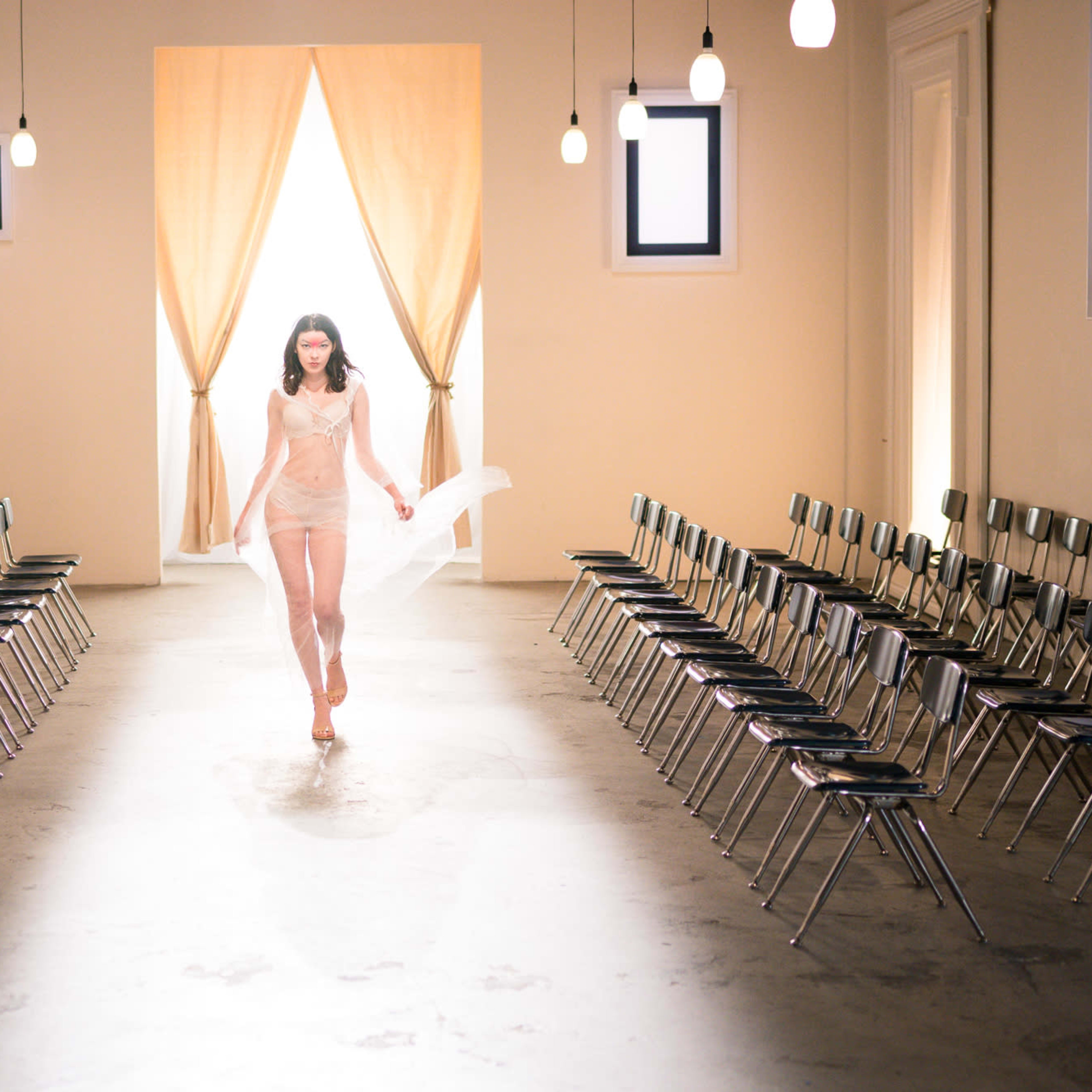 A model walks down a runway in a minimalist, well-lit space with rows of chairs on either side.