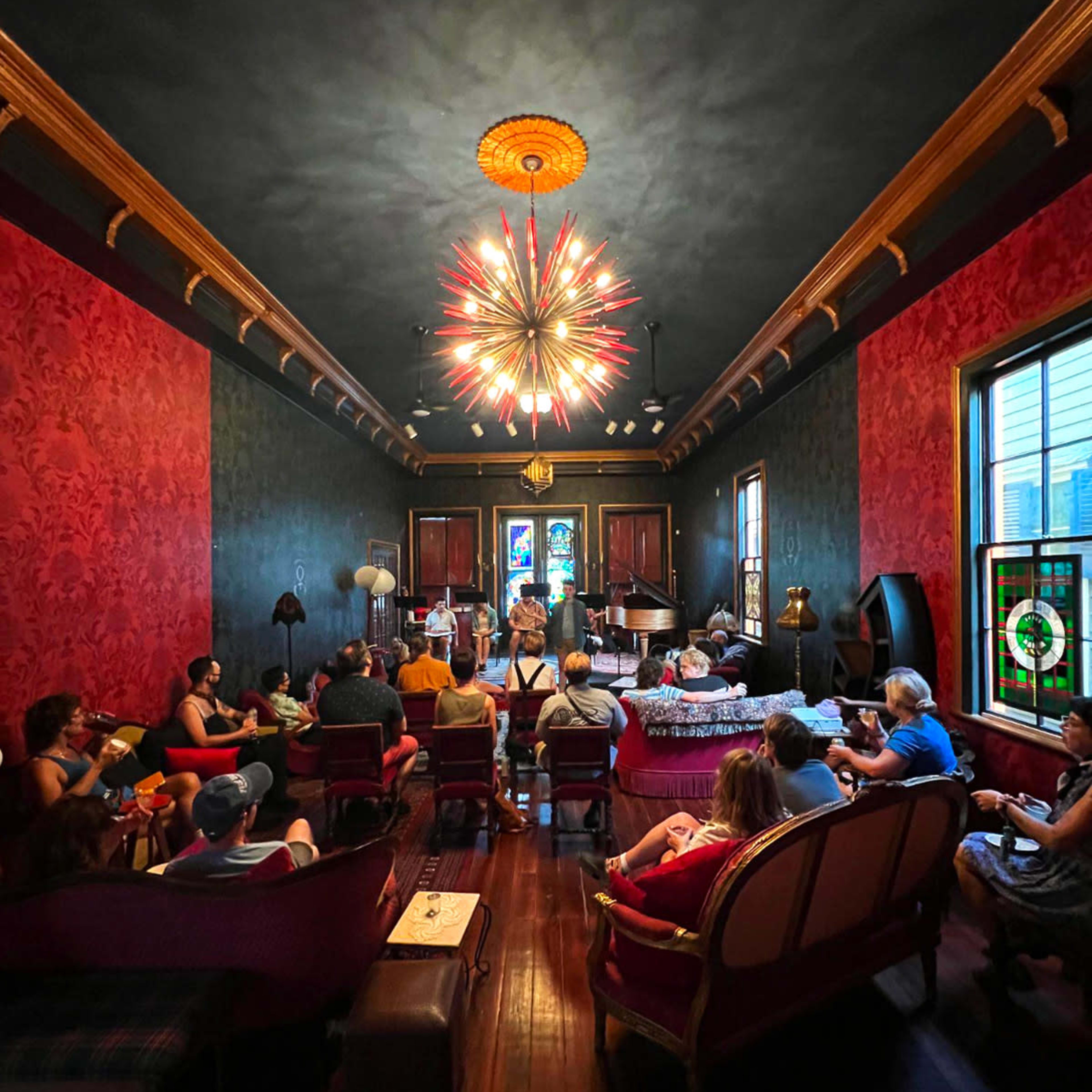 A group of people sit in a vintage-style room with patterned wallpaper and a large, modern chandelier, focused on a performer in front of them.