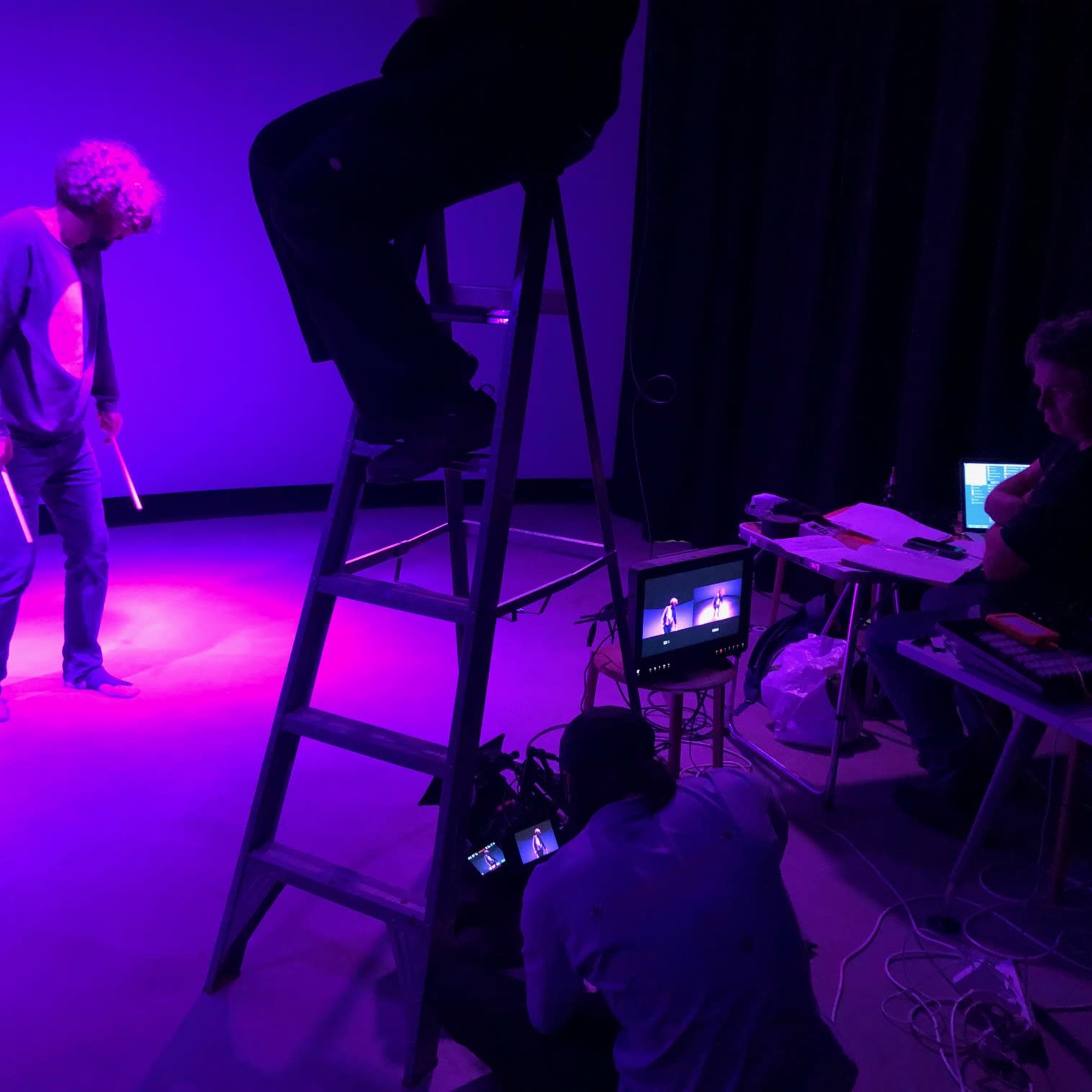 A person stands under purple lighting while another is seated at a table with equipment, and a third individual is perched on a ladder.