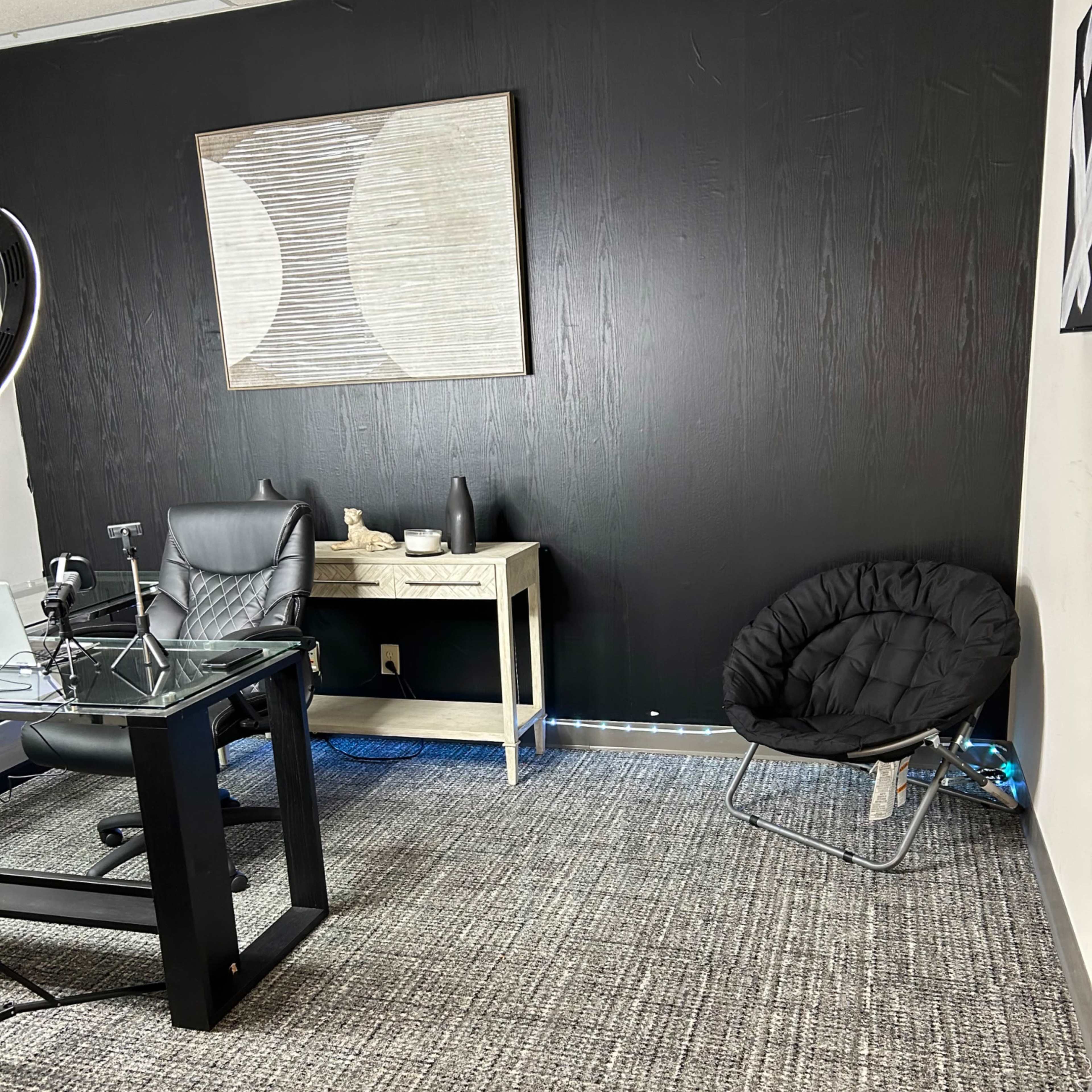 A modern office space featuring a black wall, a glass desk with a black ergonomic chair, and a round black chair in the corner.