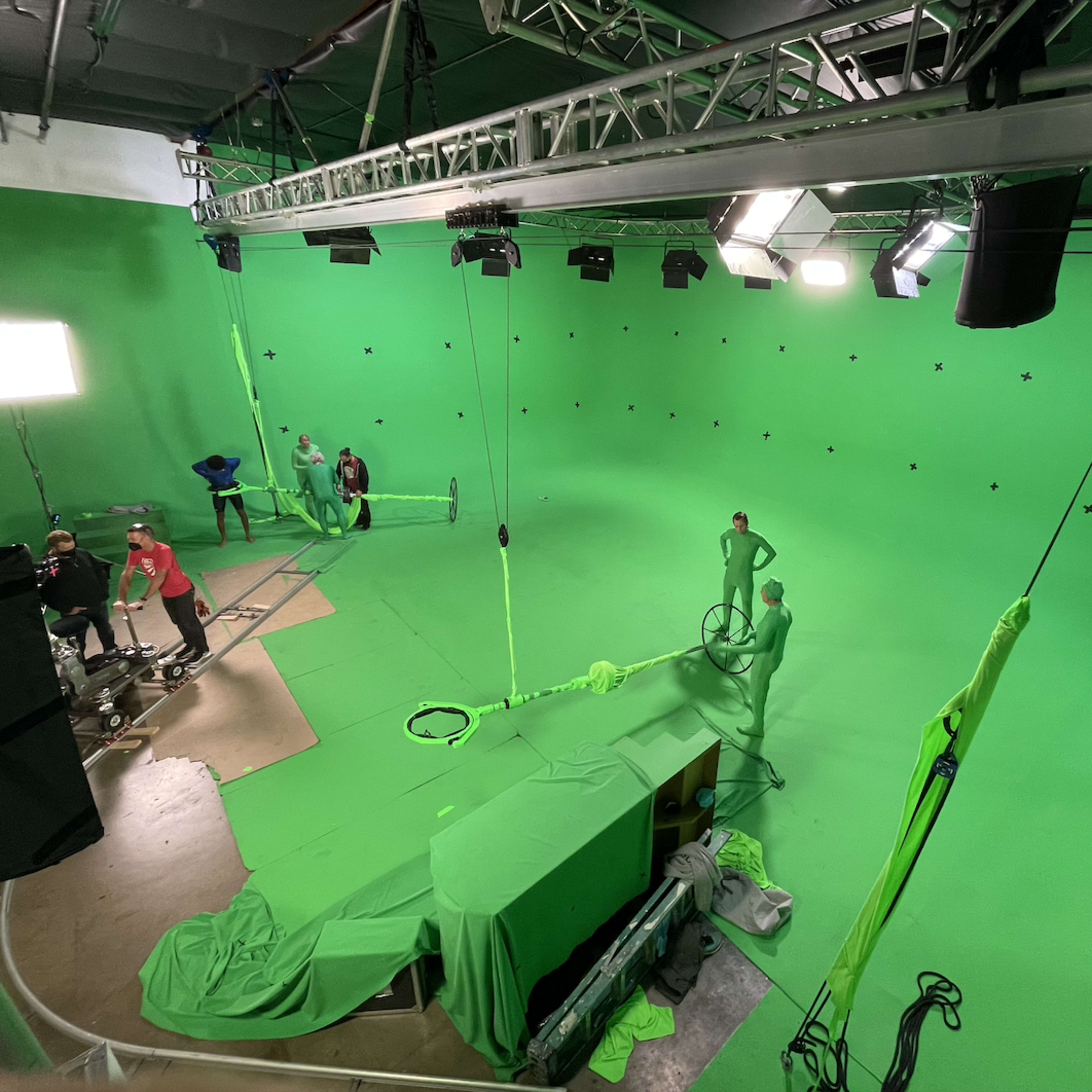 A green screen studio is set up with several crew members preparing equipment and a performer dressed in a green suit in the center.