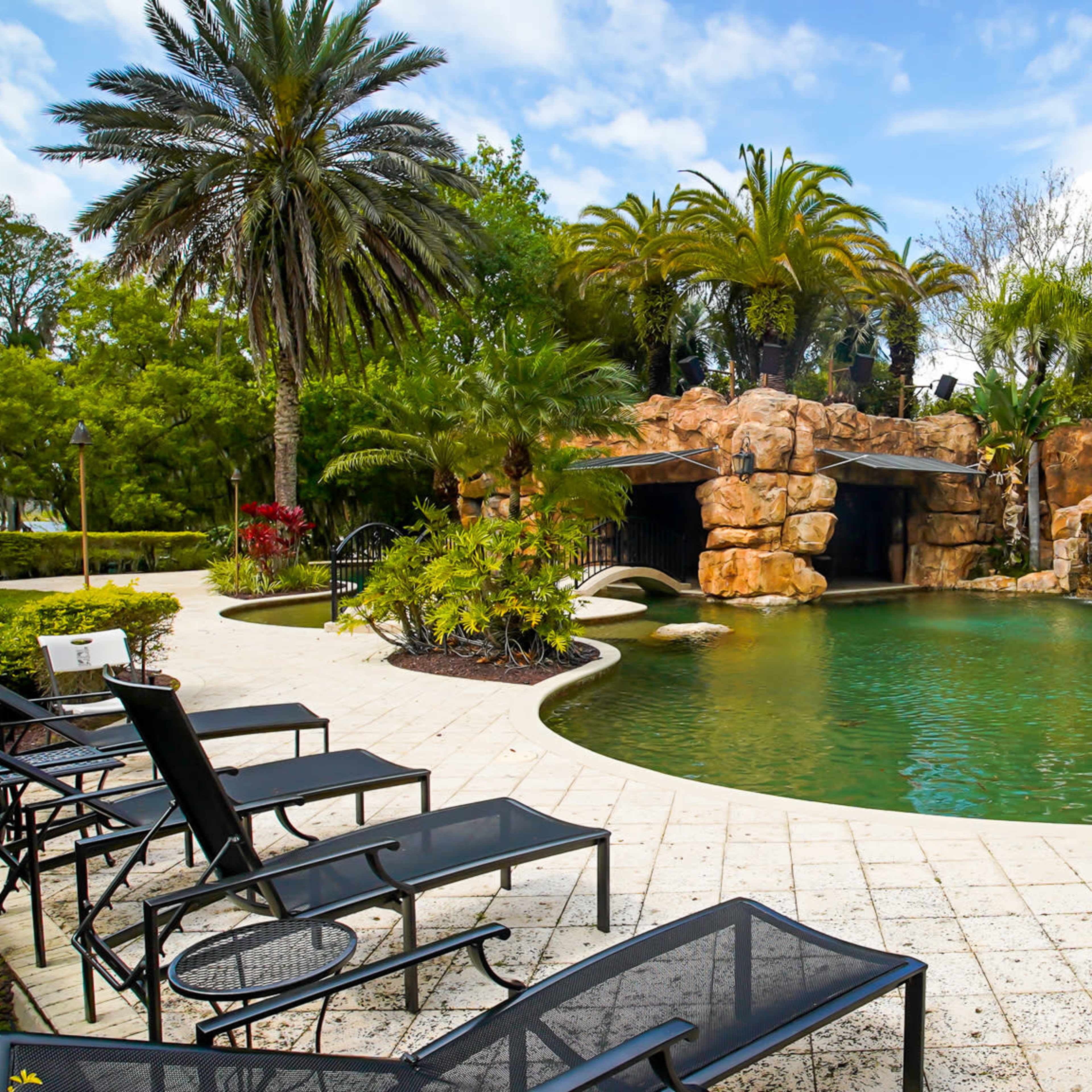 A tropical pool area features a large rock formation with water slides, surrounded by palm trees and lounge chairs.