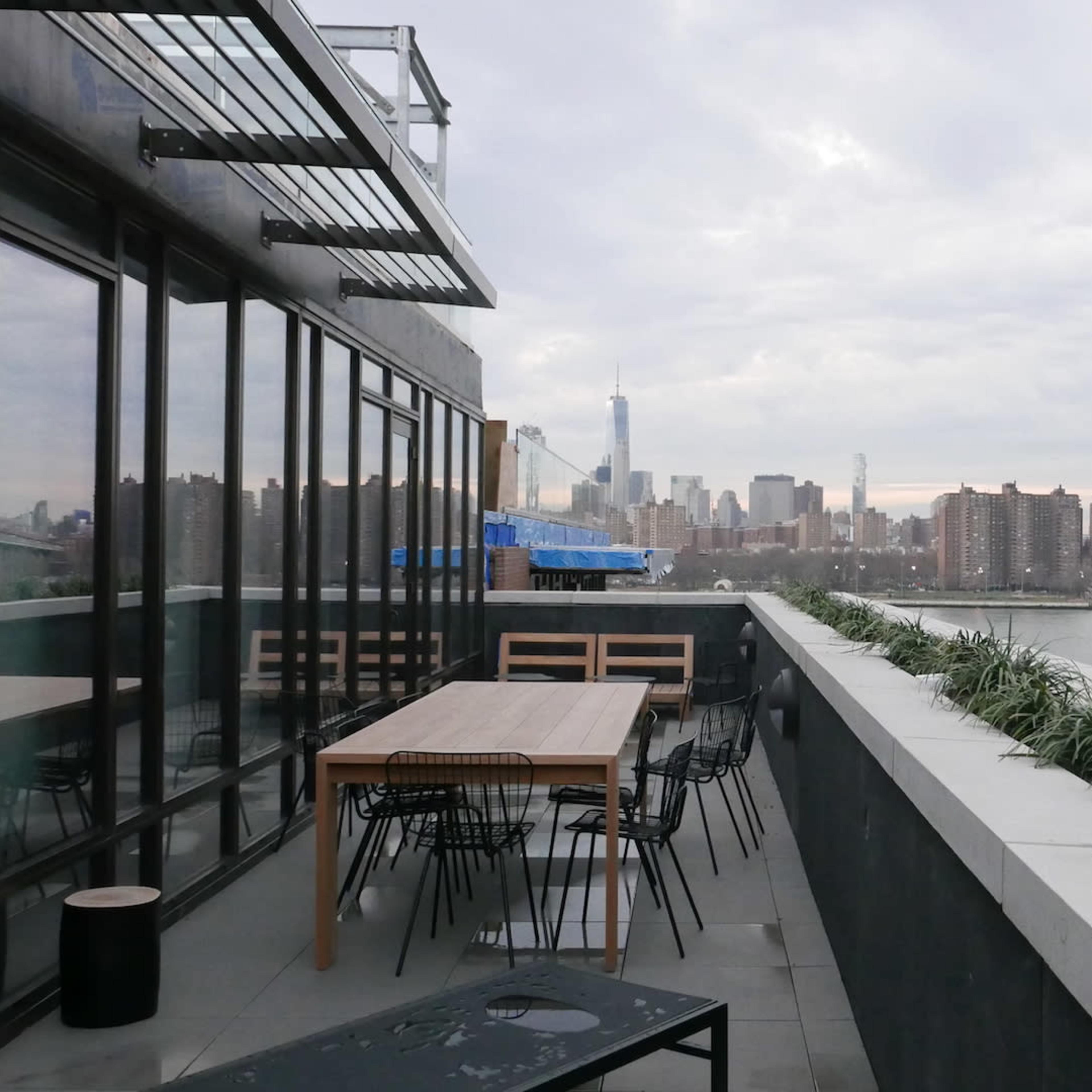 A rooftop terrace with a long table and chairs overlooking a river and a city skyline in the distance.