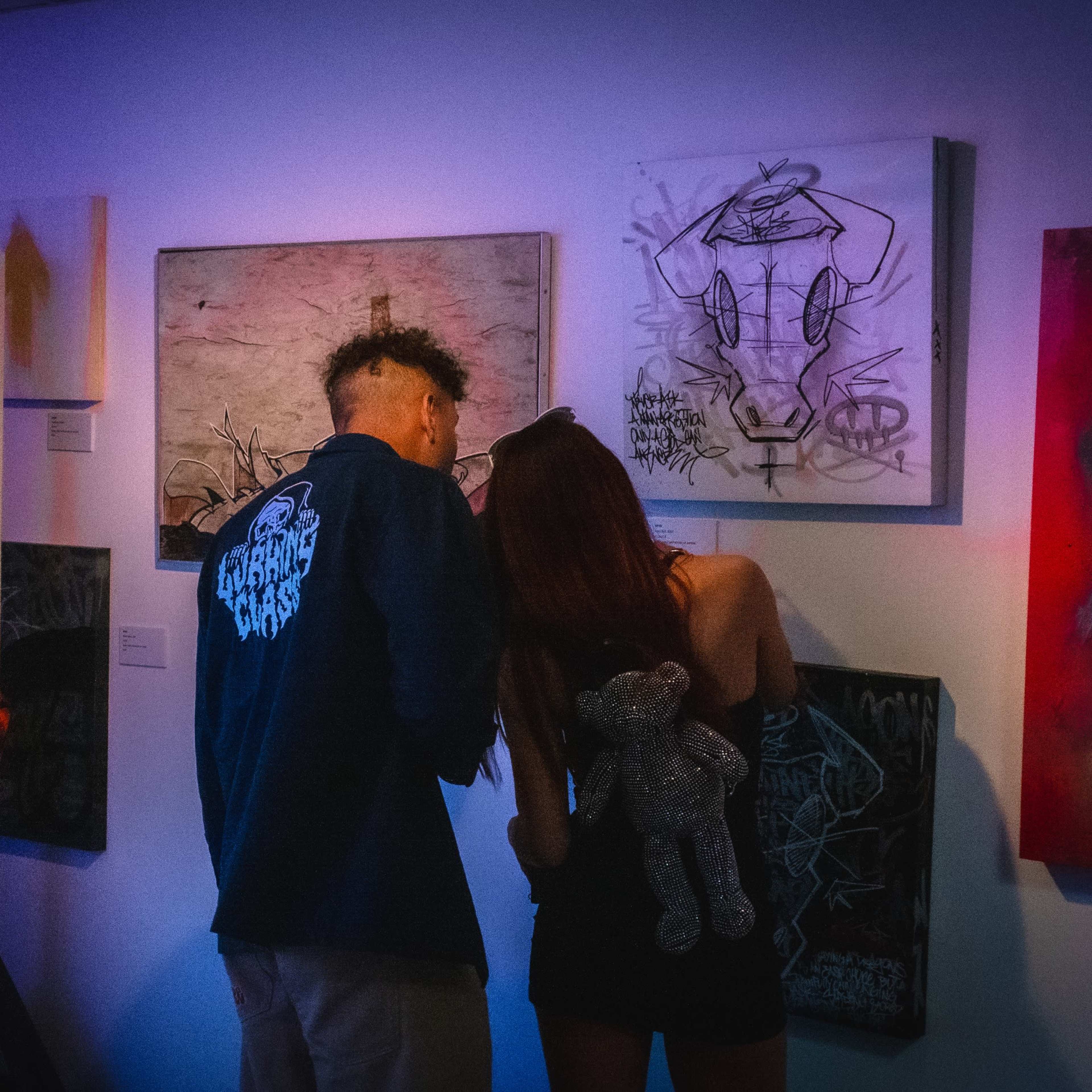 Two people examine artwork on the wall in a gallery, while a projection of a weapon is displayed on a nearby surface.