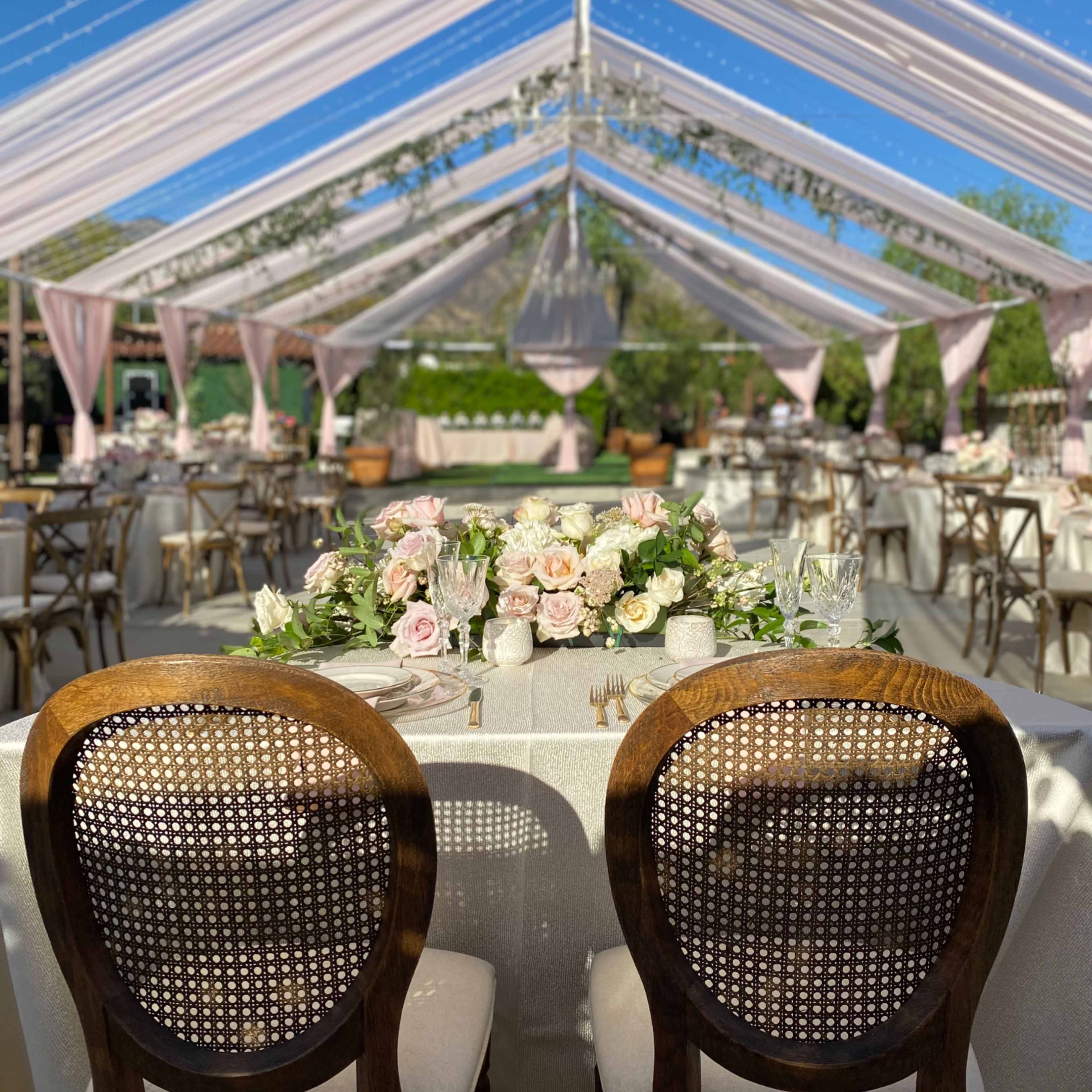 A spacious outdoor canopy tent is set up for an elegant event with decorated tables and floral arrangements.