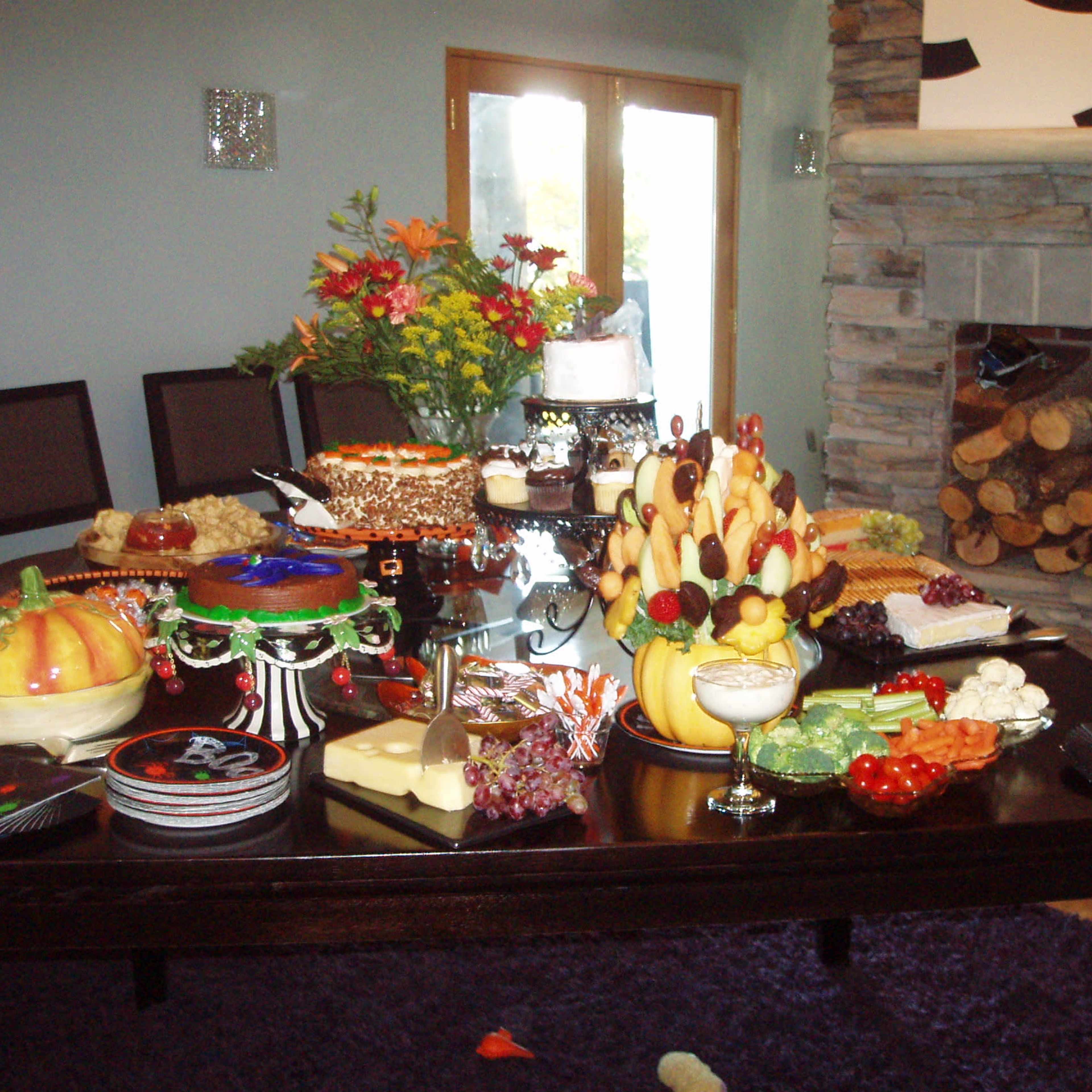 A large dining table is set with various food items, including cakes, fruits, and a decorative centerpiece of flowers, with a stone fireplace in the background.