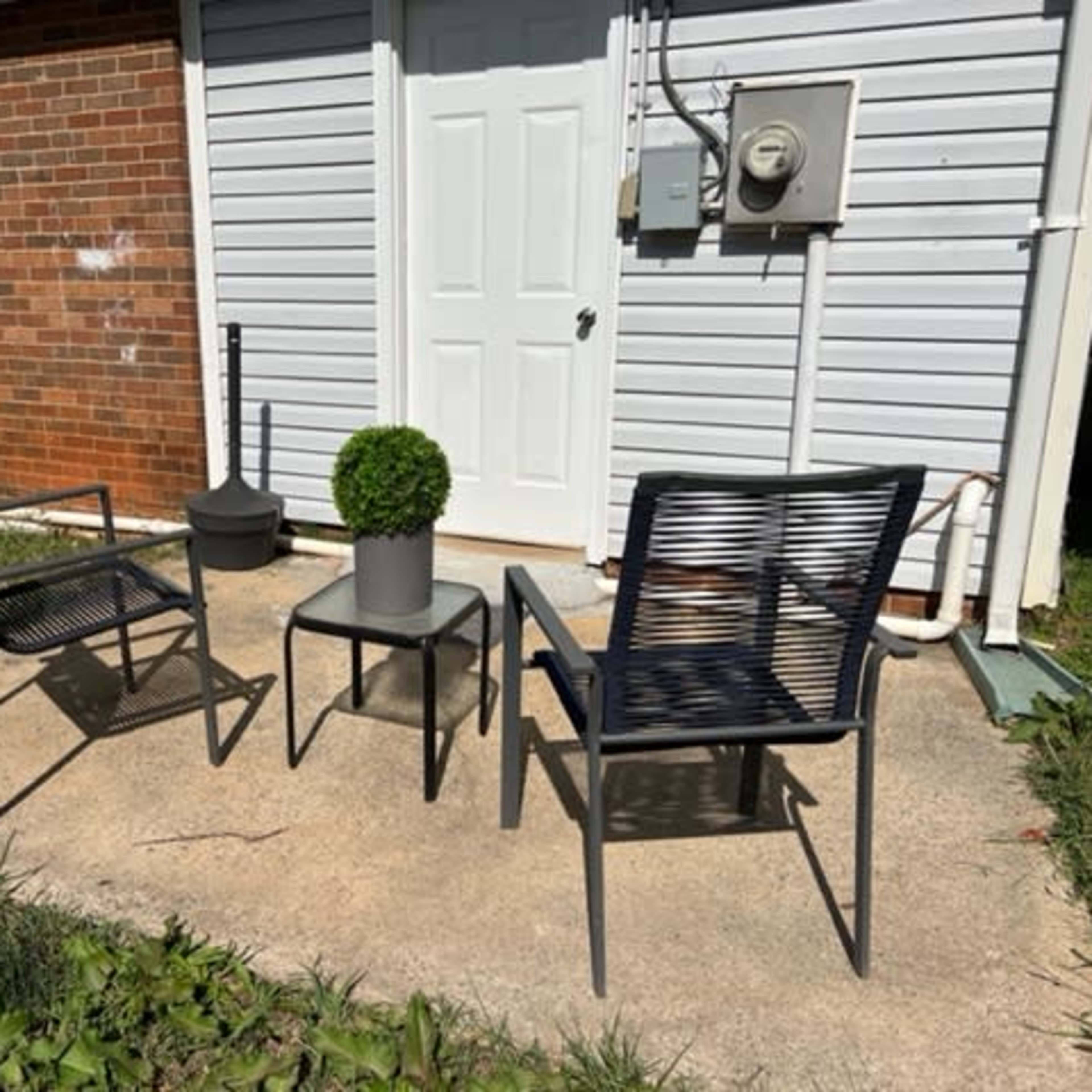 Two black chairs and a small table with a potted plant sit on a concrete patio next to a brick wall.