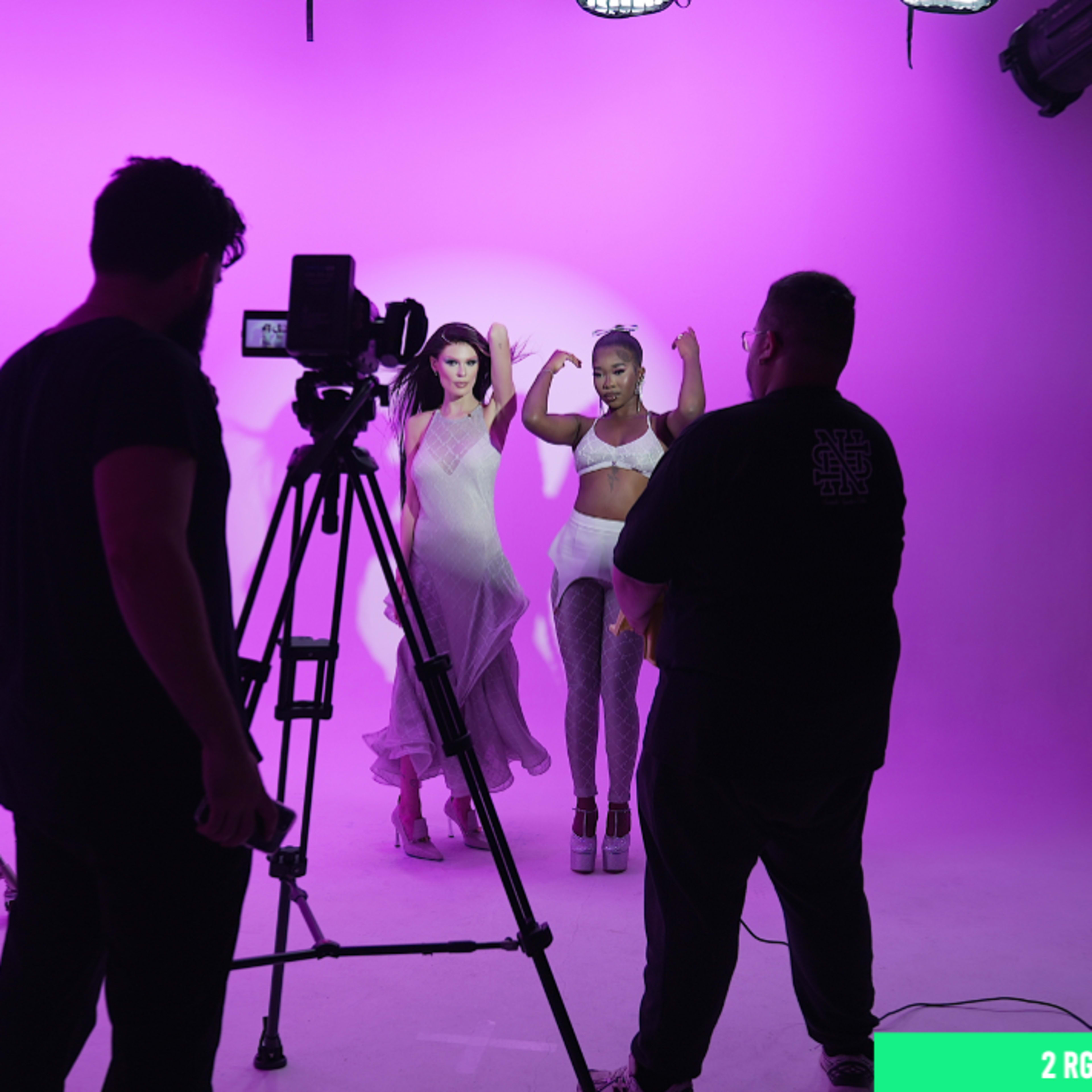 Two models pose in white outfits against a pink backdrop while a photographer sets up a camera and additional lighting equipment.
