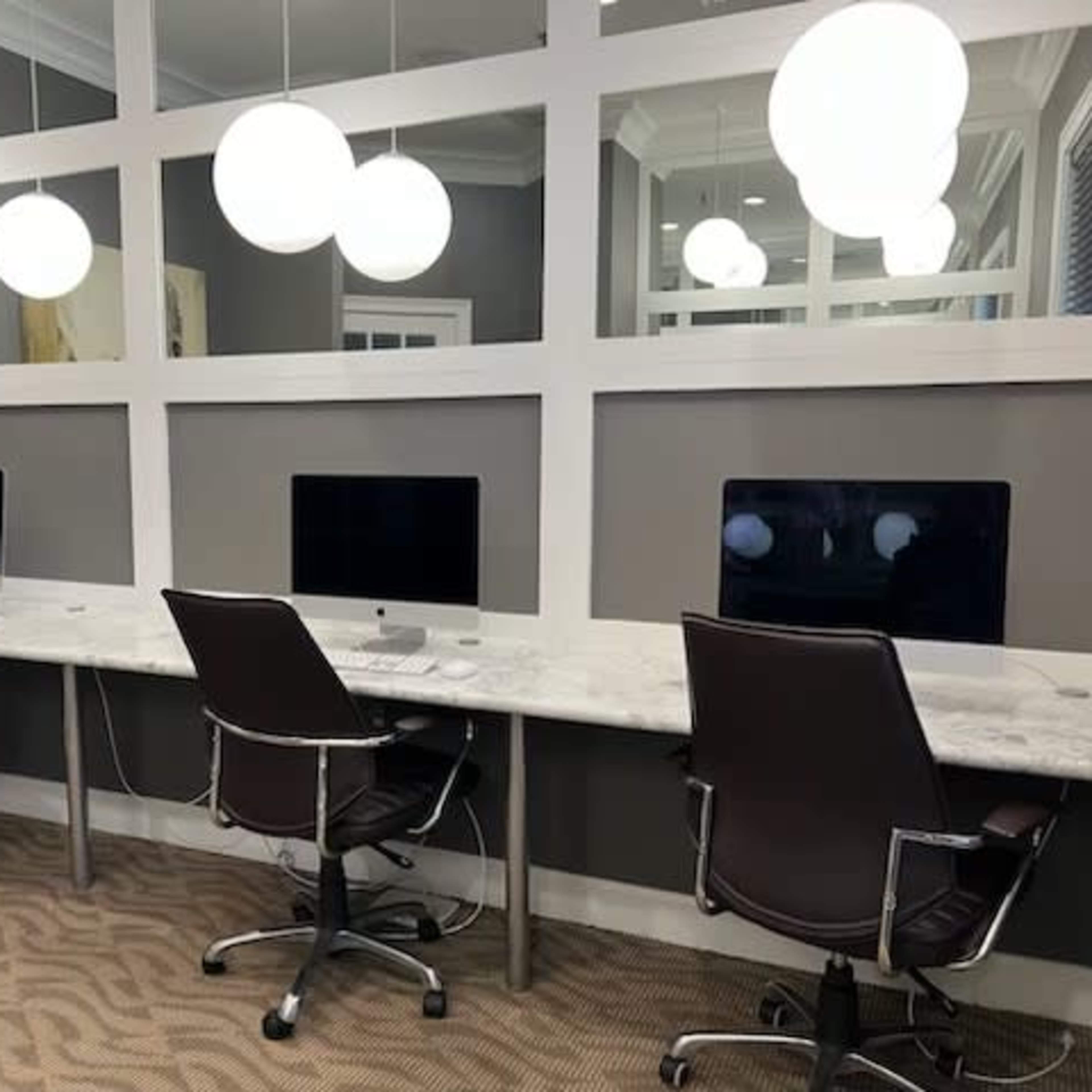 A modern workspace with three desktop computers on marble-top desks, each accompanied by swivel chairs and illuminated by round pendant lights above.