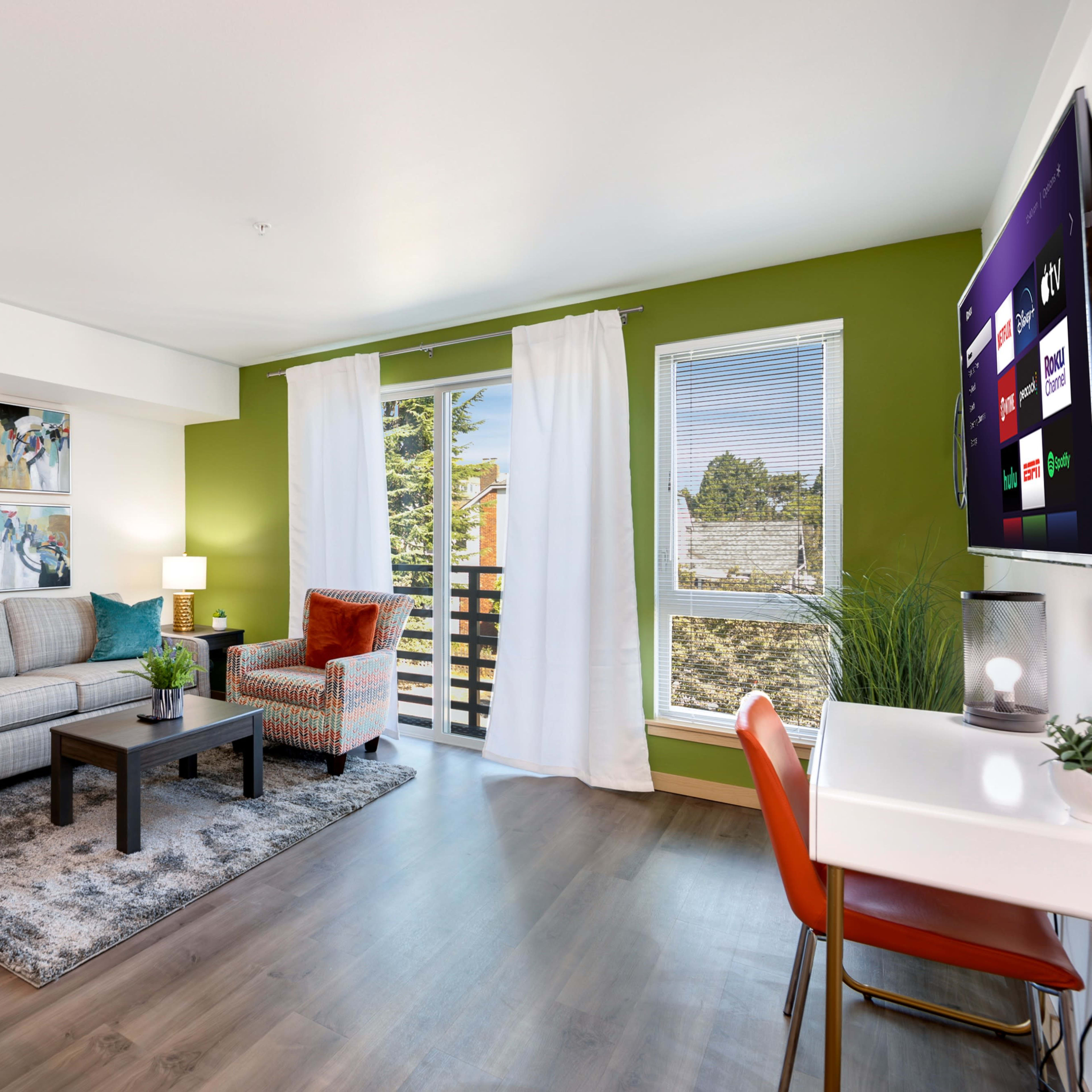 A modern living room with a sofa, coffee table, and a large TV, featuring green walls and large windows that open to a balcony.