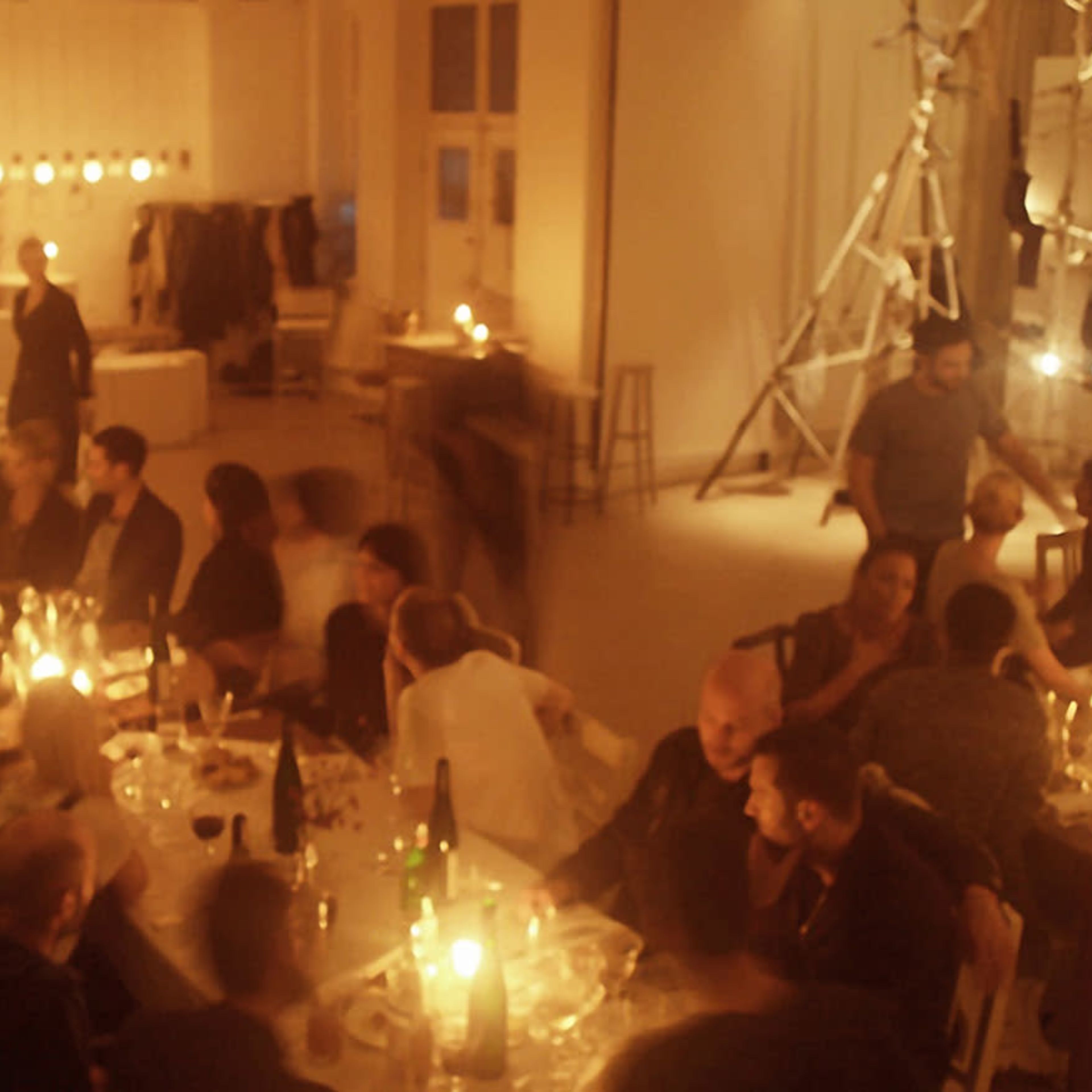 A dimly lit dining space is filled with people gathered around long tables adorned with candles and plates during a festive gathering.