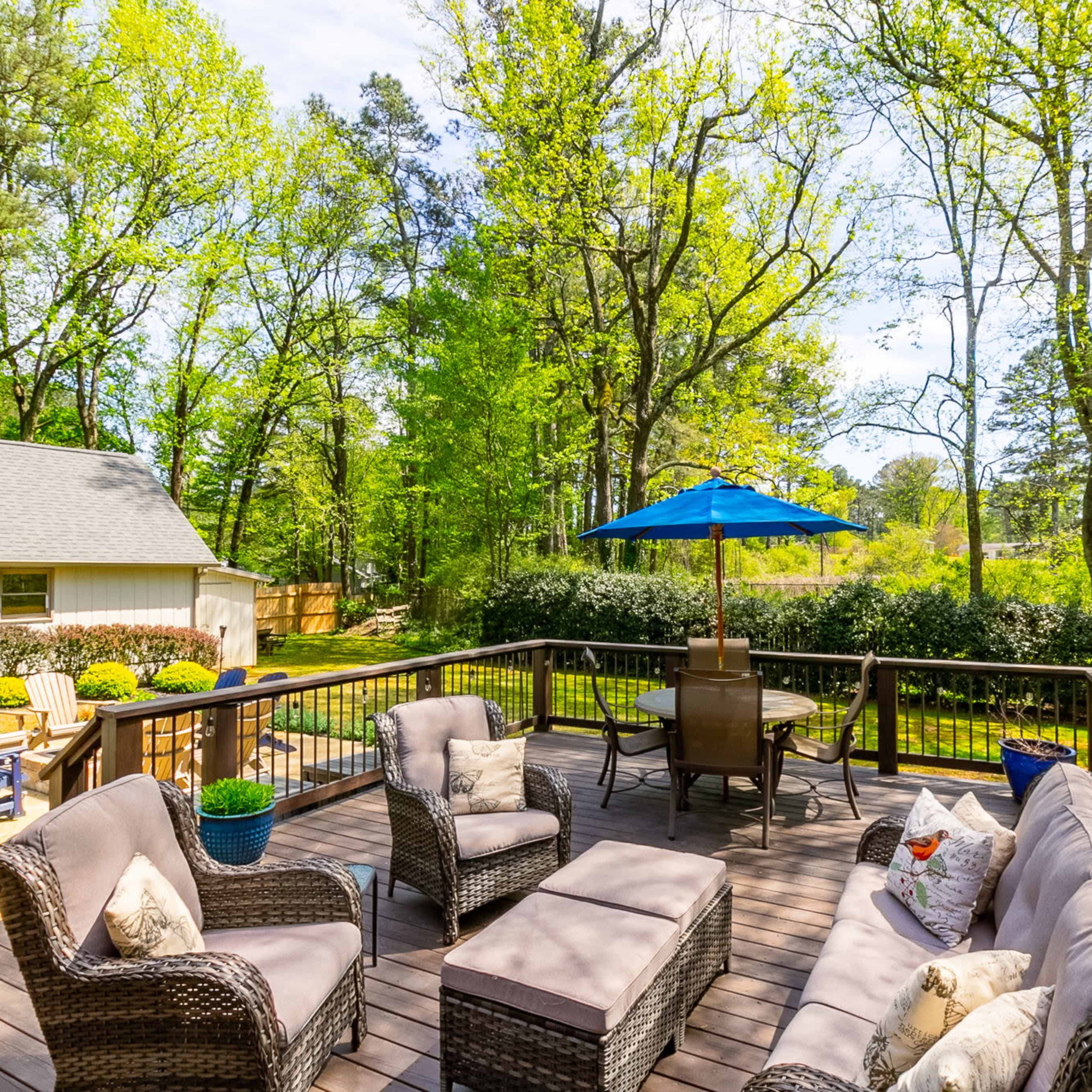 A wooden deck features outdoor seating with a blue umbrella and lush greenery in the background.