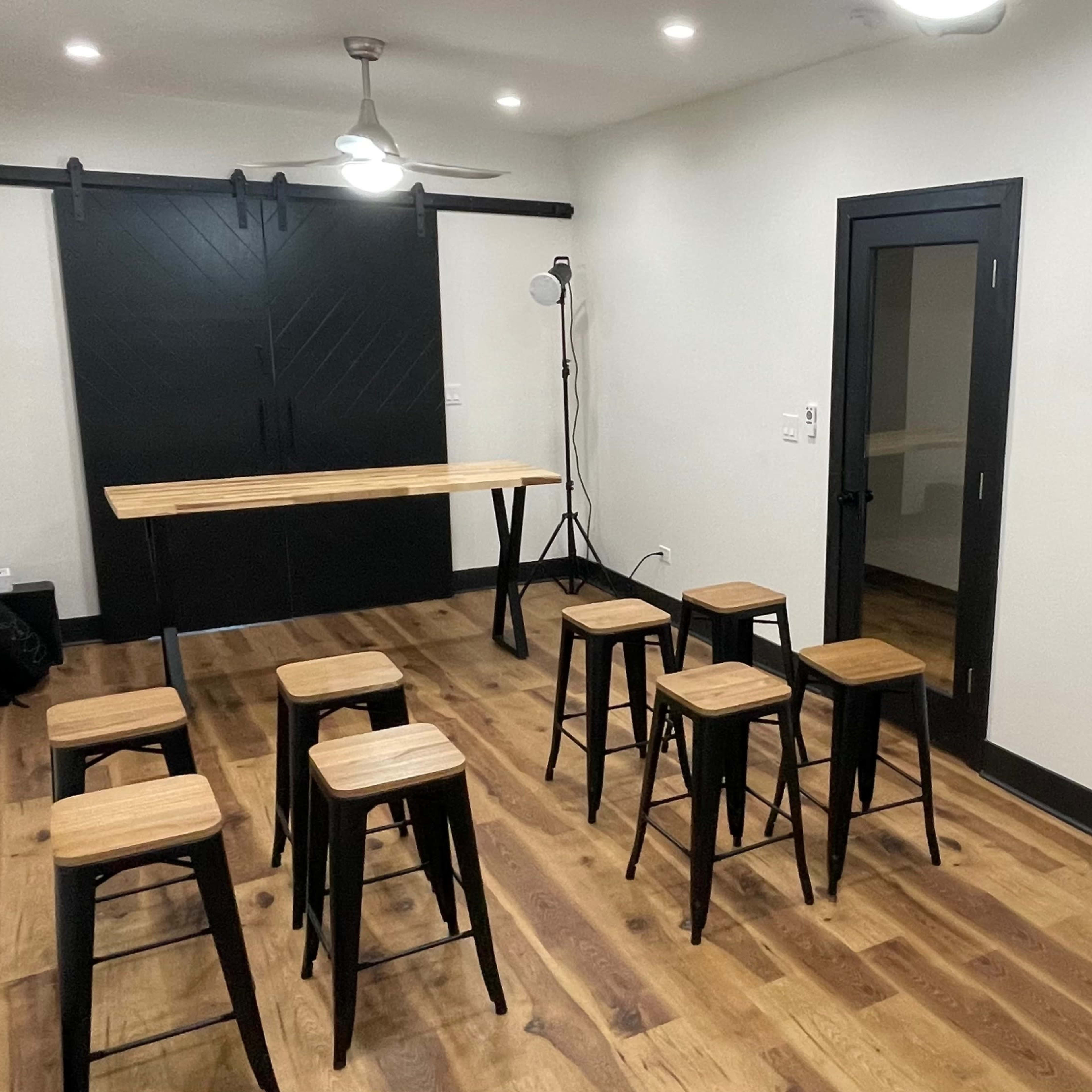 A modern meeting room features a long wooden table surrounded by eight black stools, with a wall-mounted TV and sliding barn door.