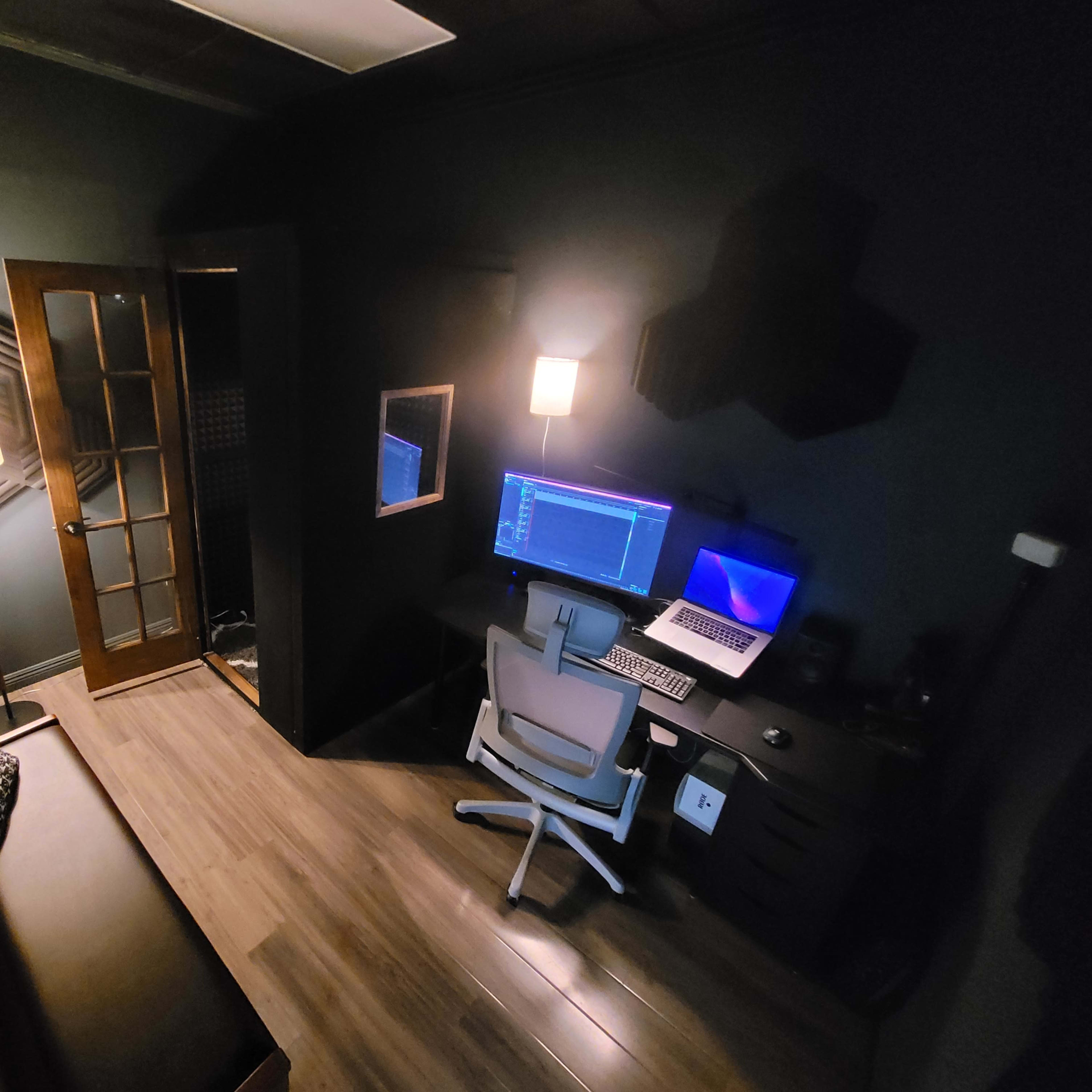 A dimly lit room featuring a desk with a computer, a chair, a lamp, and a door leading to another space.
