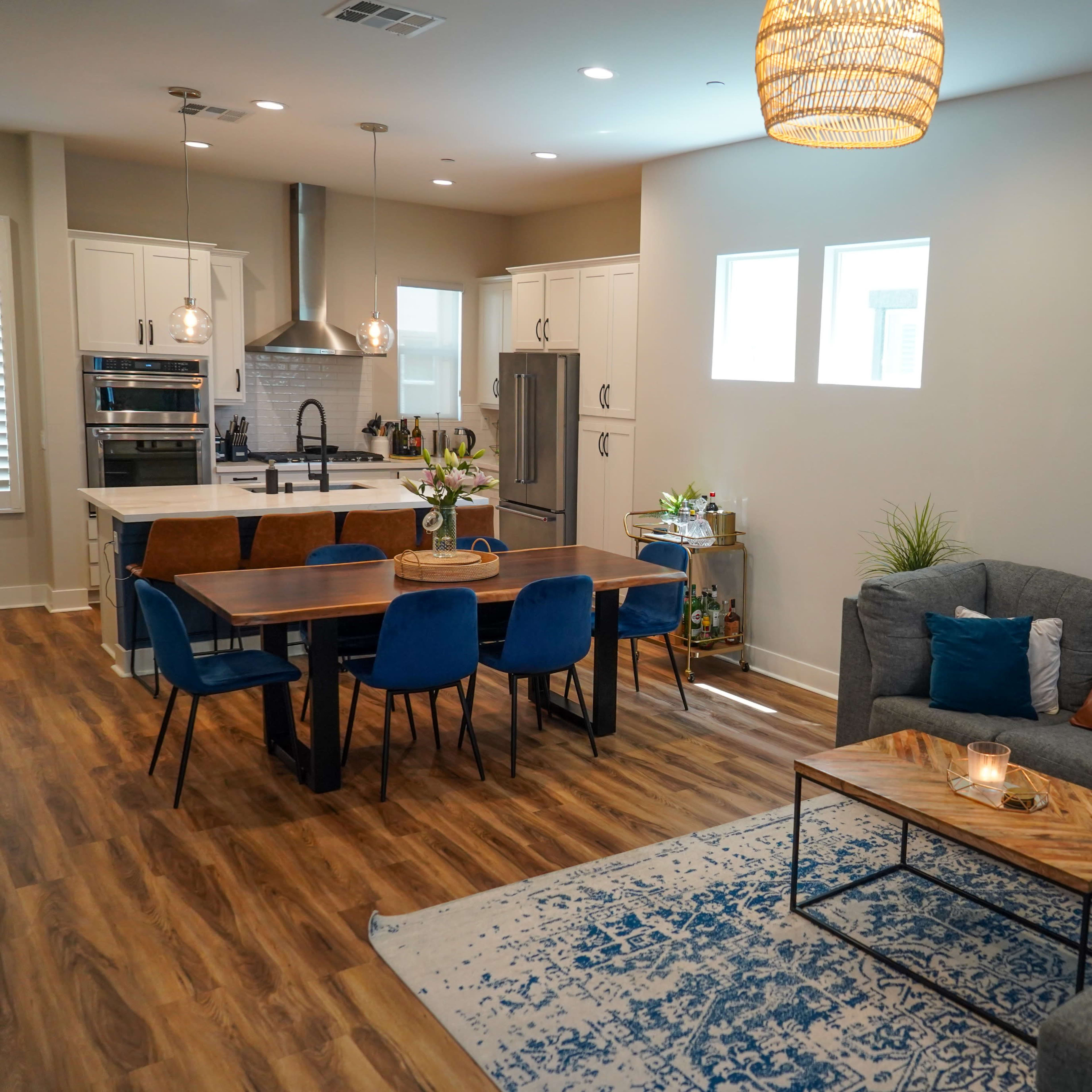A modern living and dining area features a gray sectional sofa, a wooden dining table with blue chairs, and a kitchen in the background.