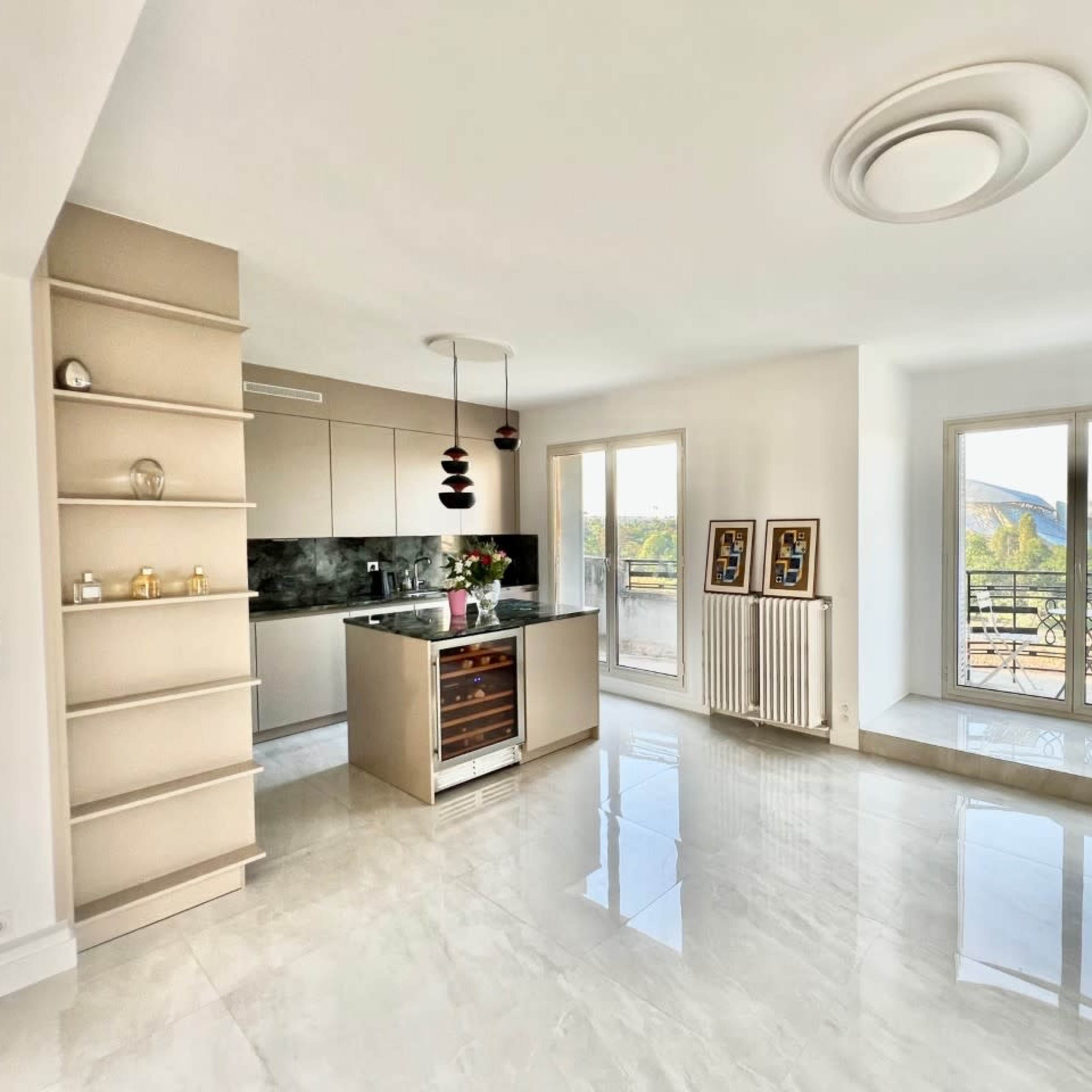 A modern, open-concept living area with a kitchen, a shelf unit, and large windows overlooking a balcony and outdoor landscape.