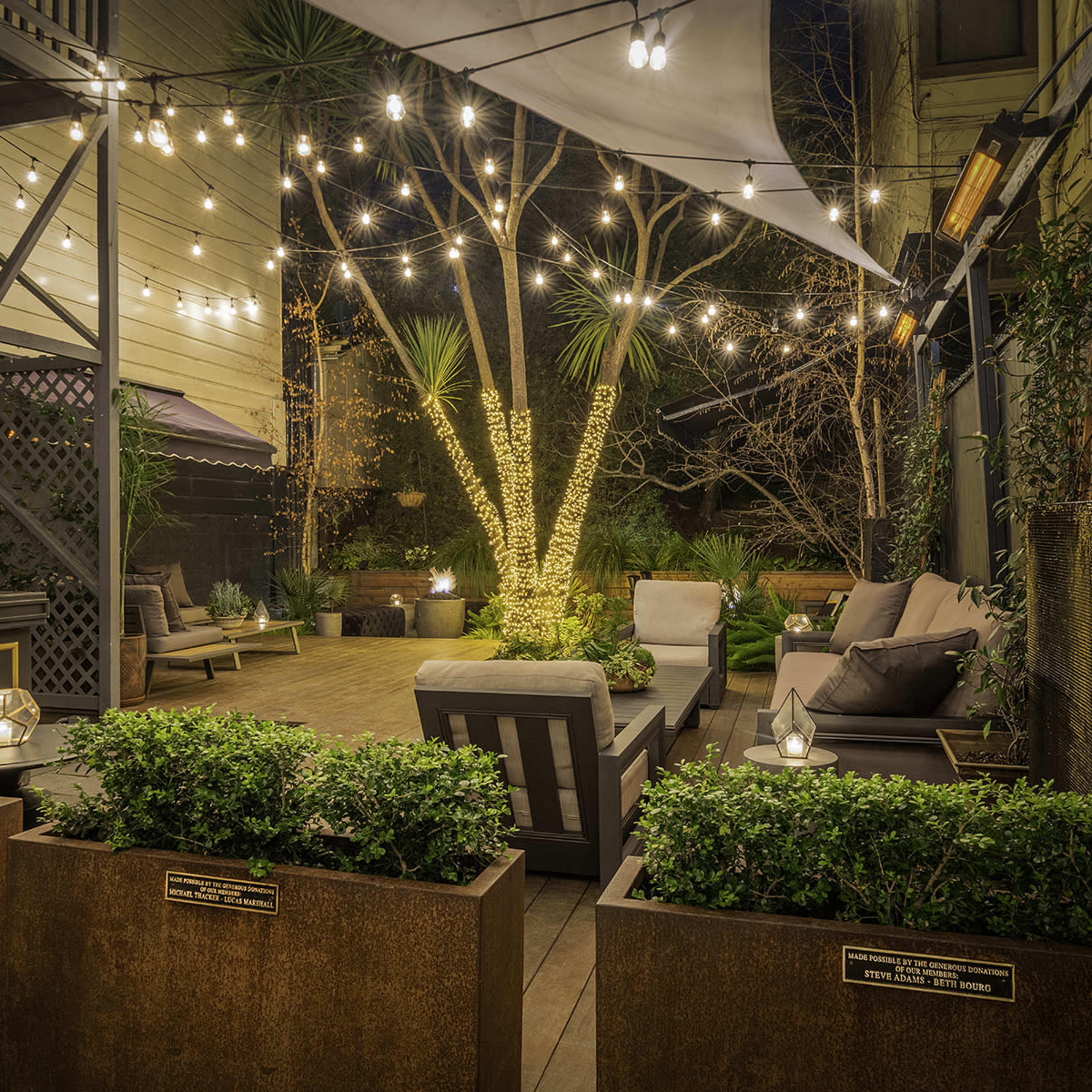 A landscaped outdoor patio with string lights, potted plants, and seating arrangements in a cozy, inviting setting.