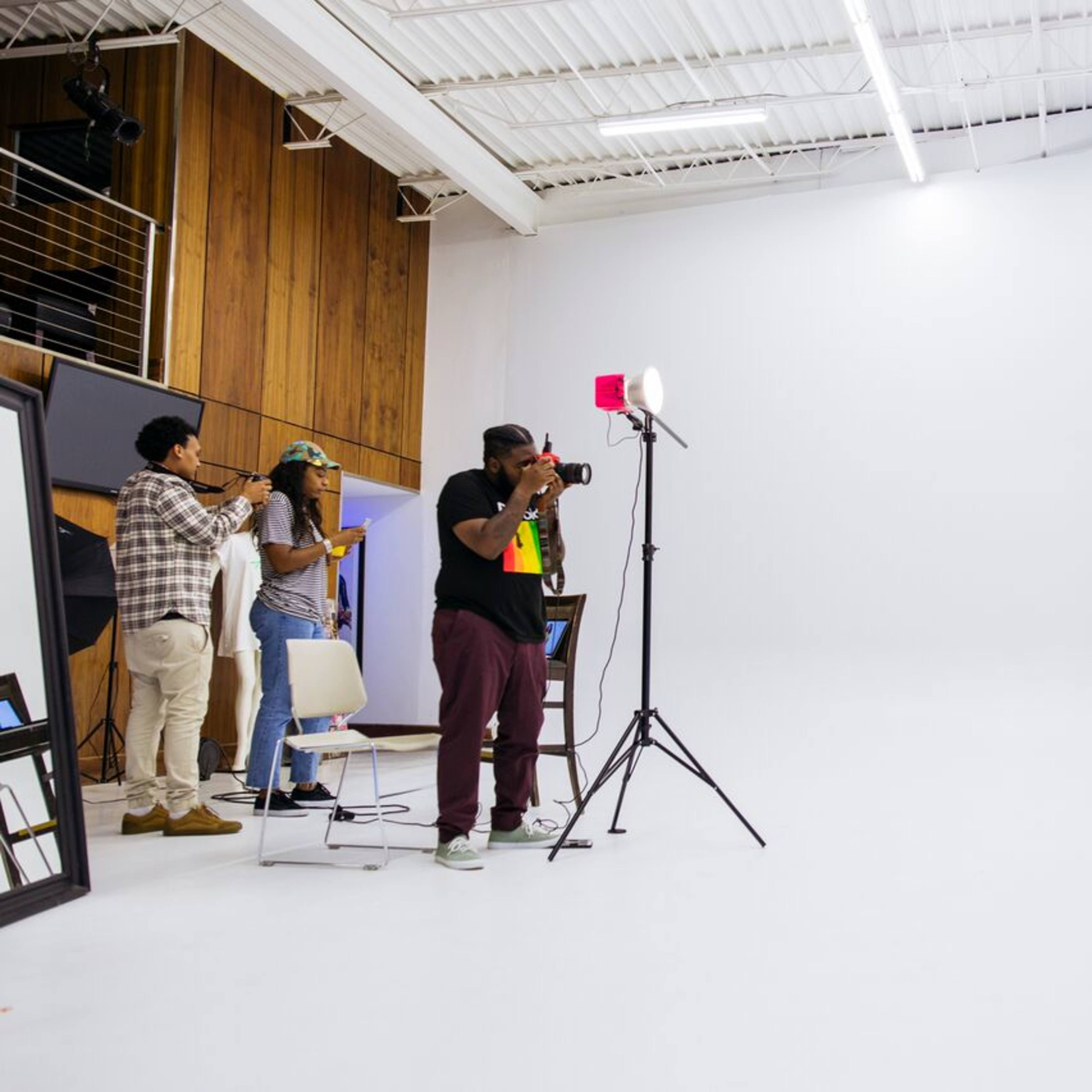 A photoshoot is taking place in a studio with a model posing against a white backdrop while photographers and crew are set up to capture the images.