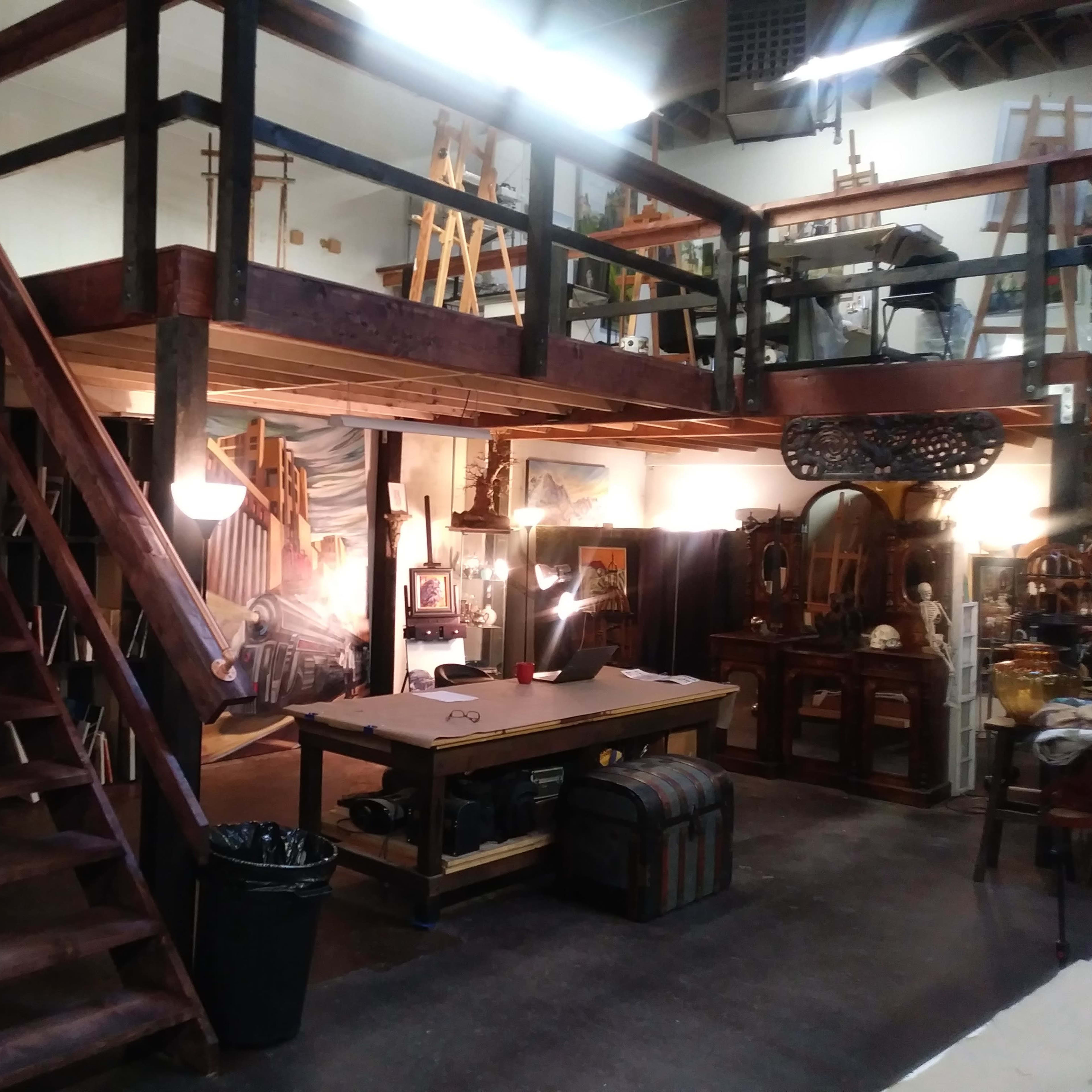 An art studio with a lofted area accessible by a wooden staircase, a central work table, and various art supplies and furniture.