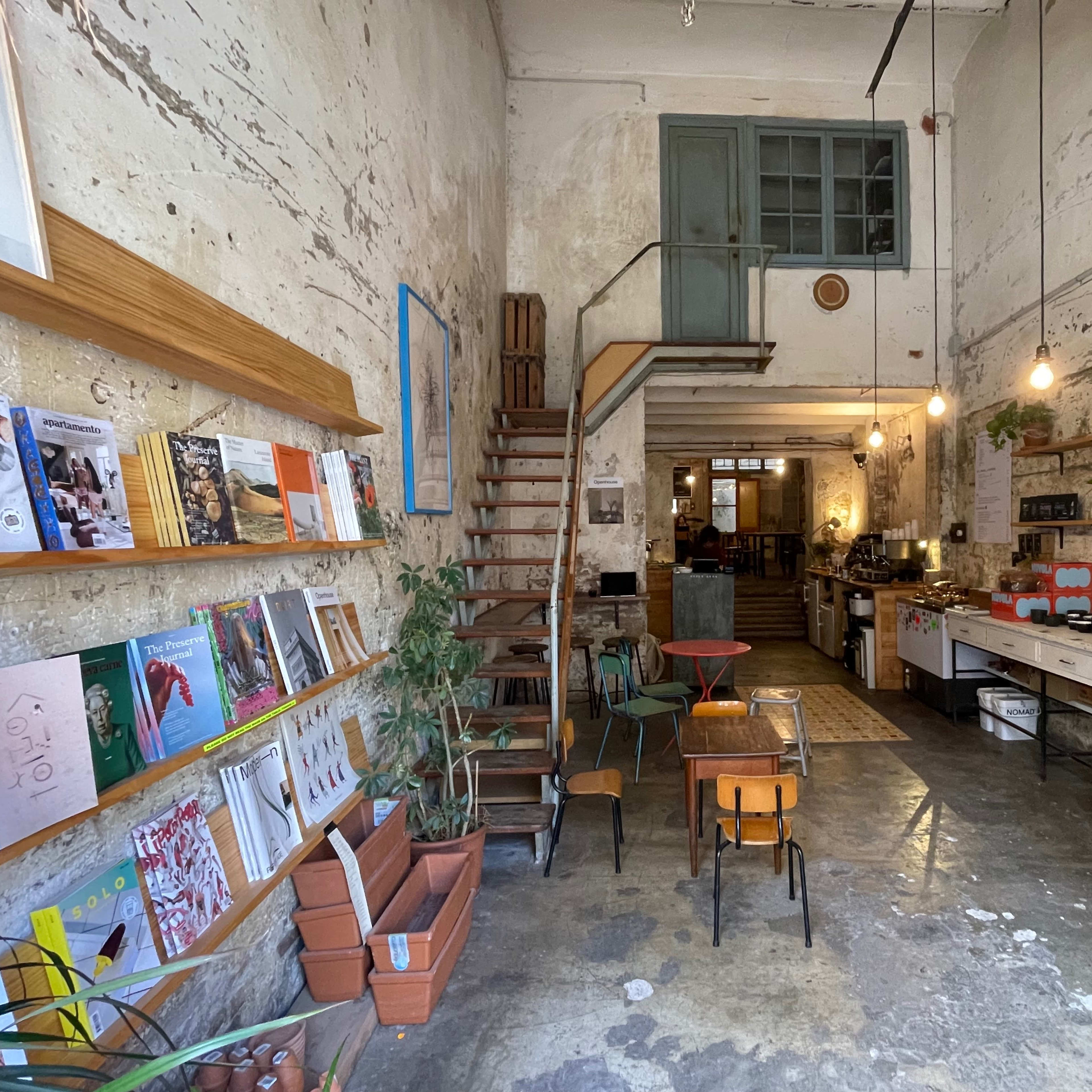A rustic café with exposed brick walls, wooden stairs leading to an upper level, and shelves filled with books and magazines.