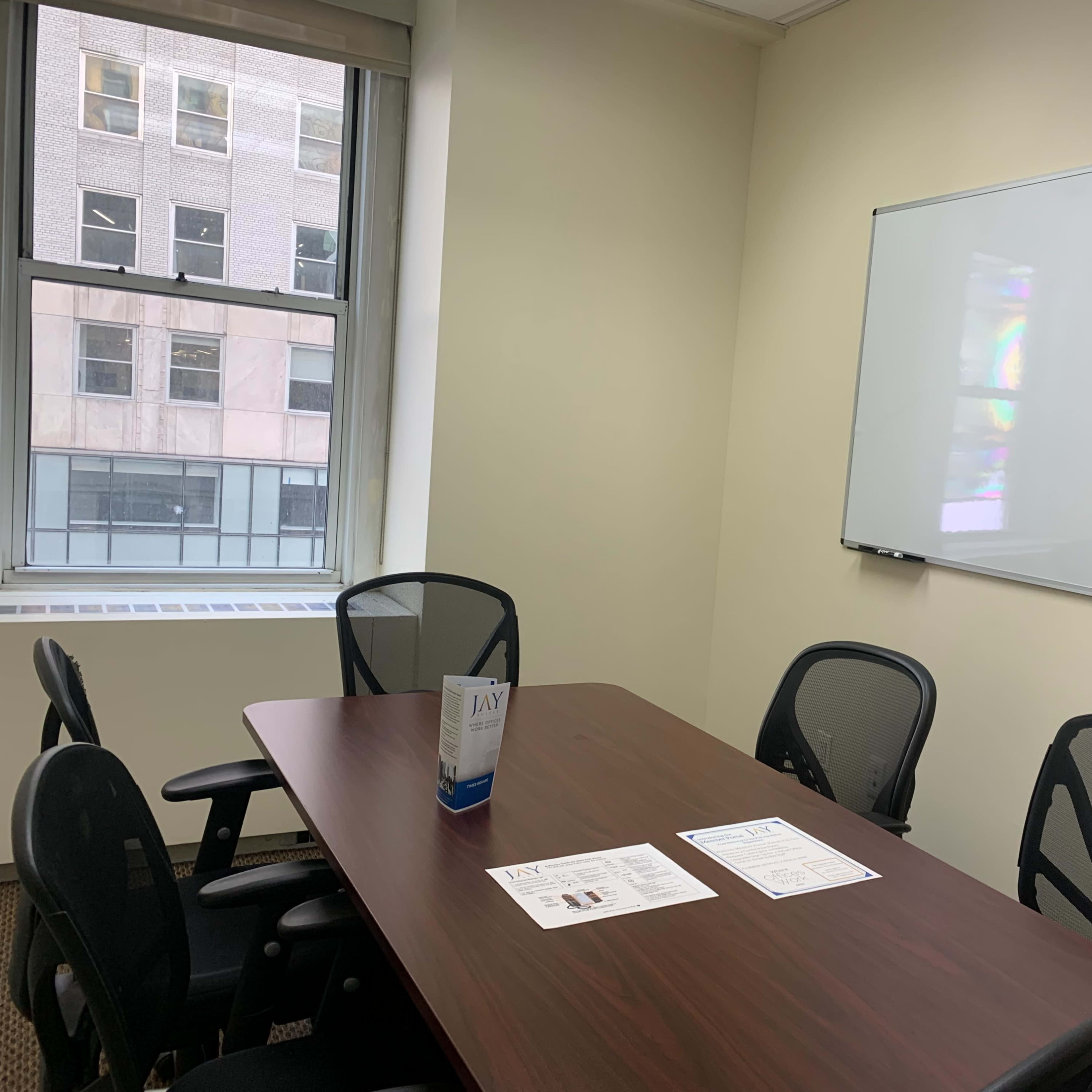 A small conference room with a wooden table, four black chairs, and a whiteboard on the wall, along with a few printed documents on the table.