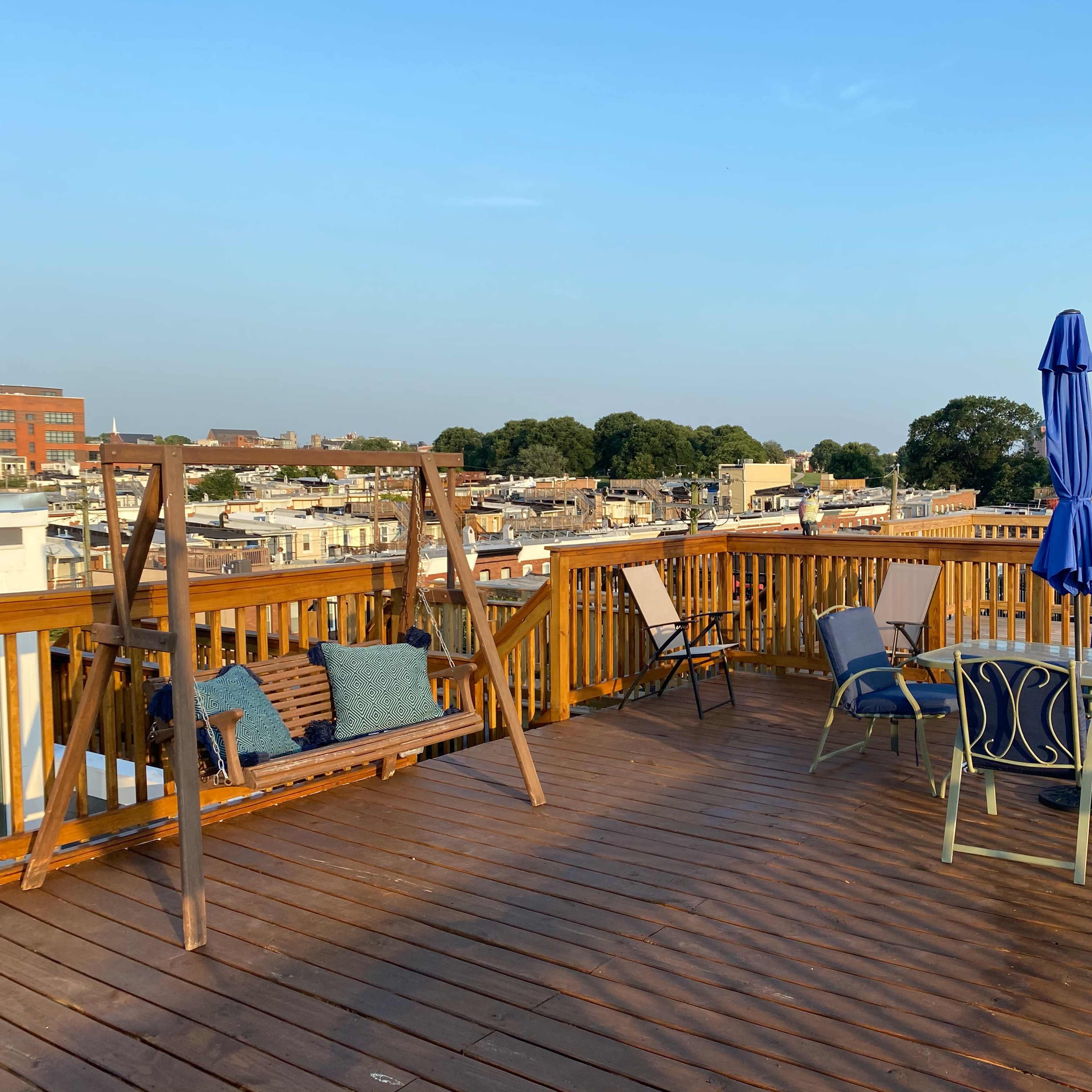 A rooftop deck with a wooden swing, several chairs, a table, and an umbrella, overlooking a cityscape.