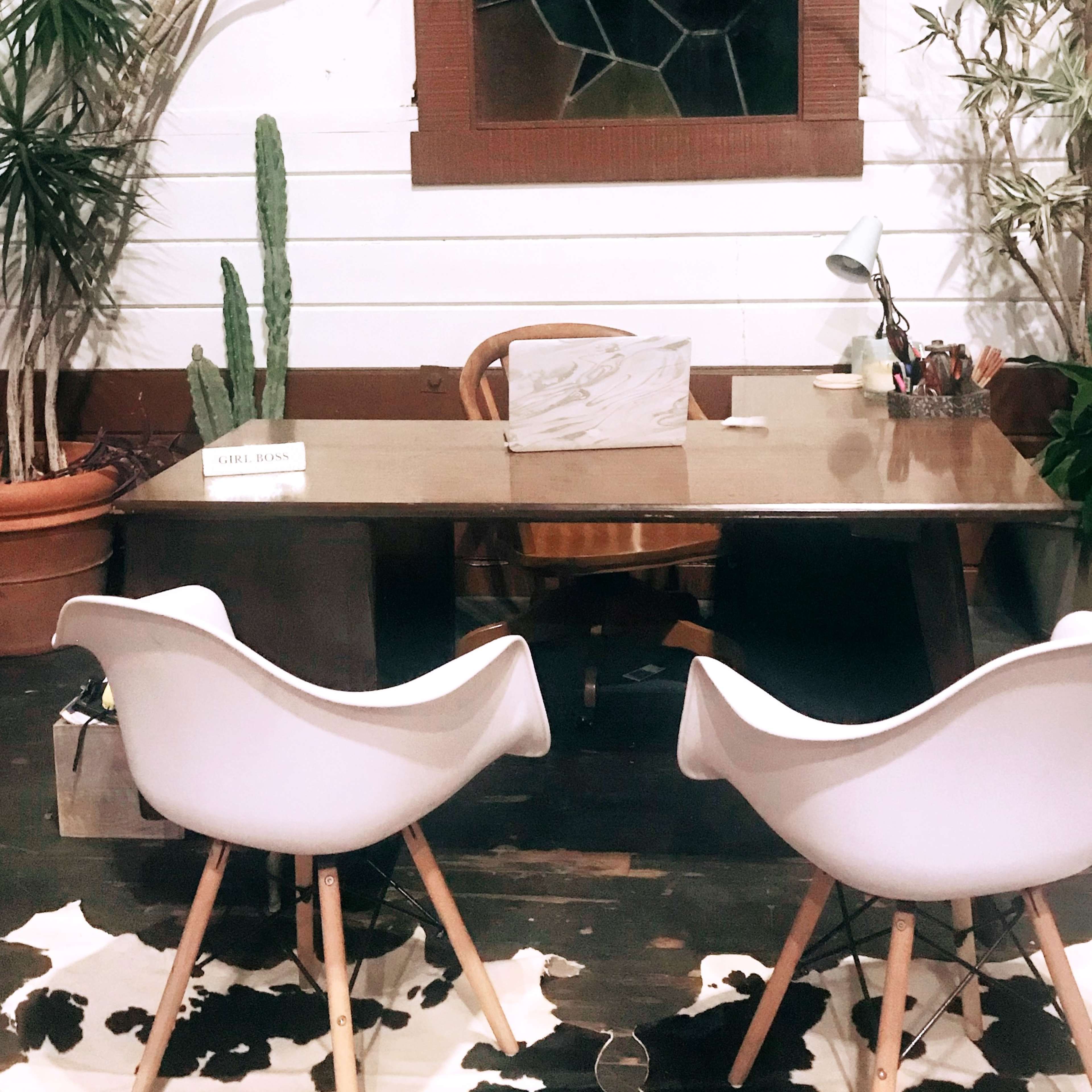 A minimalist office space featuring a wooden desk, two white chairs, and several potted plants against a wall with a stained glass window.