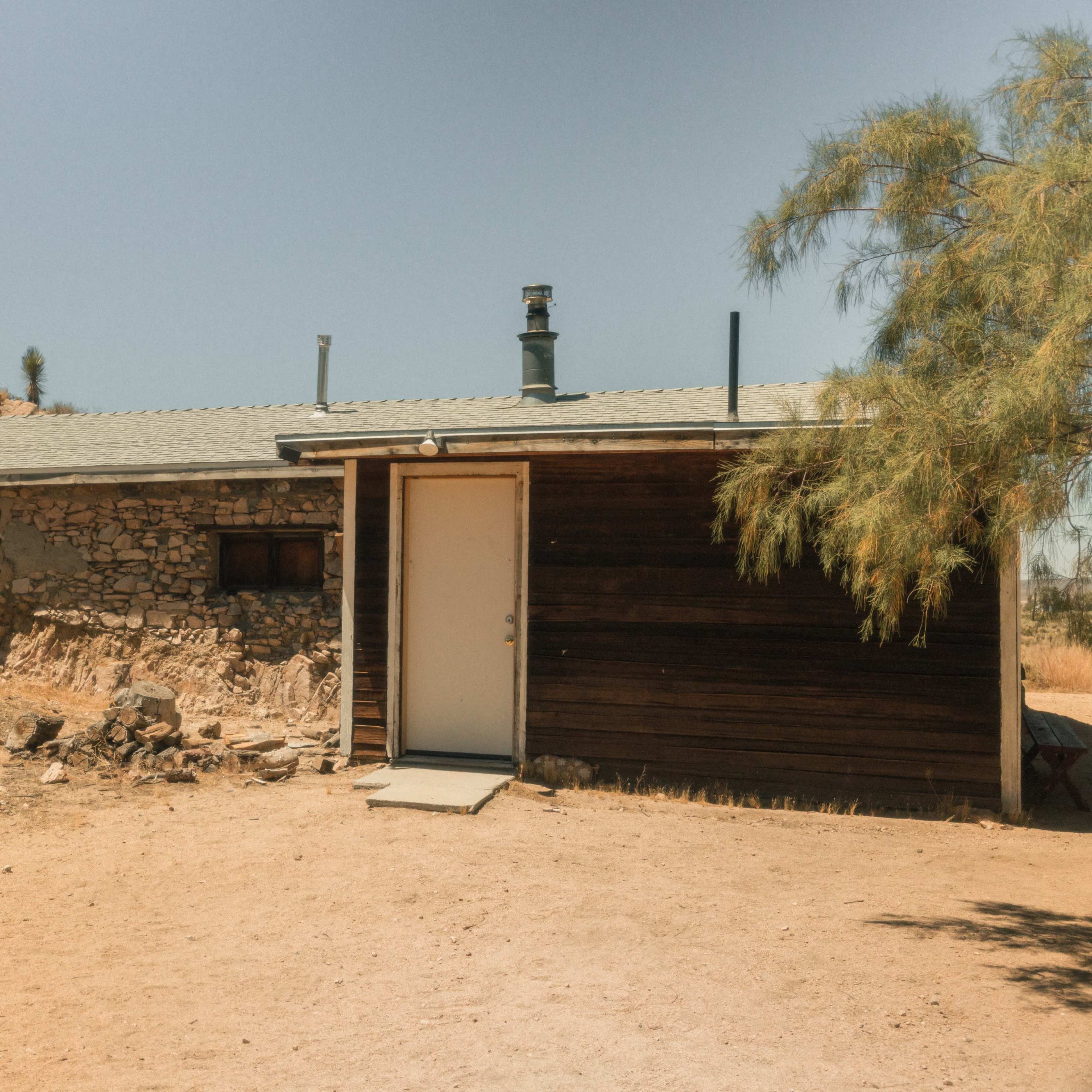 A small, rustic house with a stone wall and wooden siding is situated on a rocky, dry hillside.