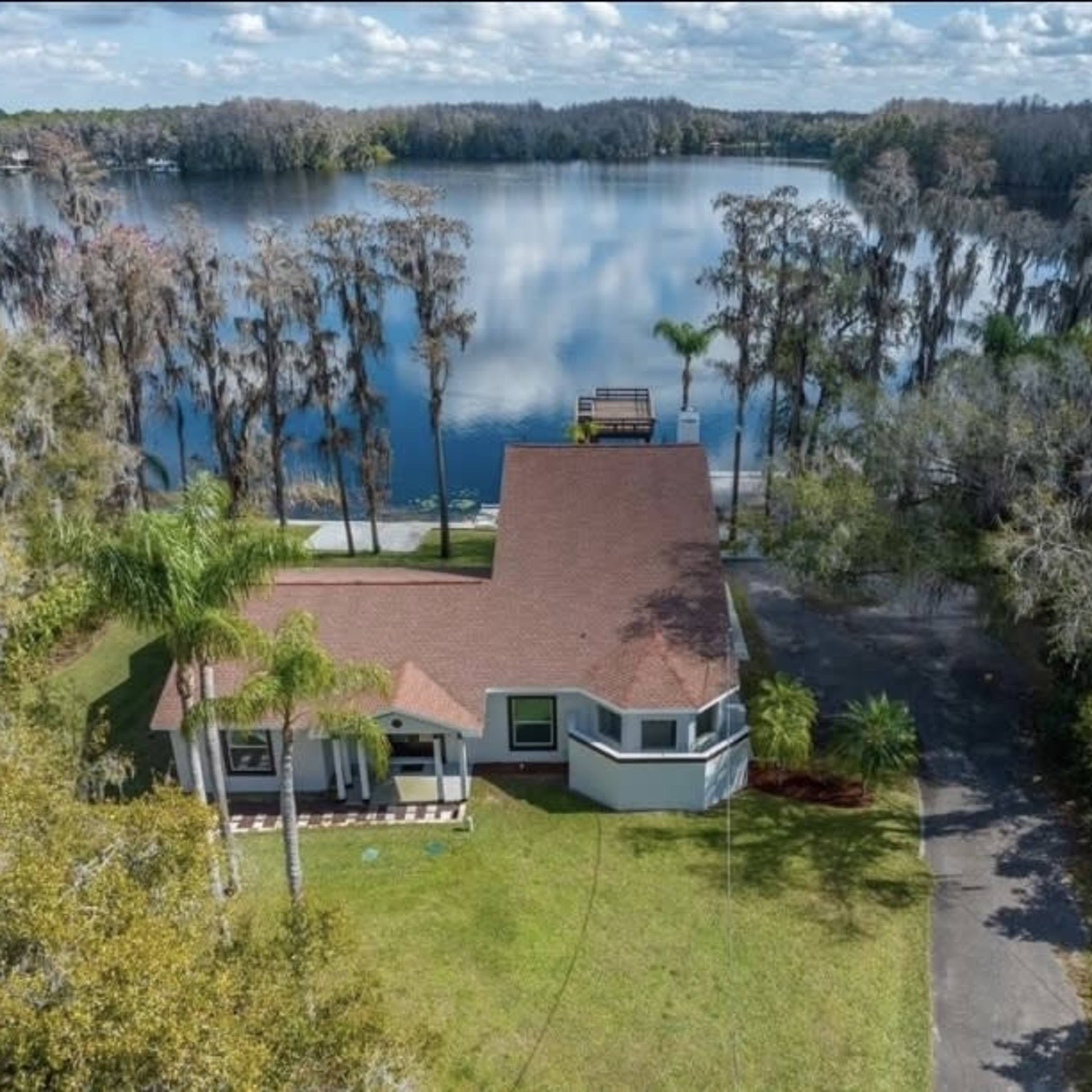 Aerial view of a single-story house with a red roof beside a calm lake surrounded by trees.