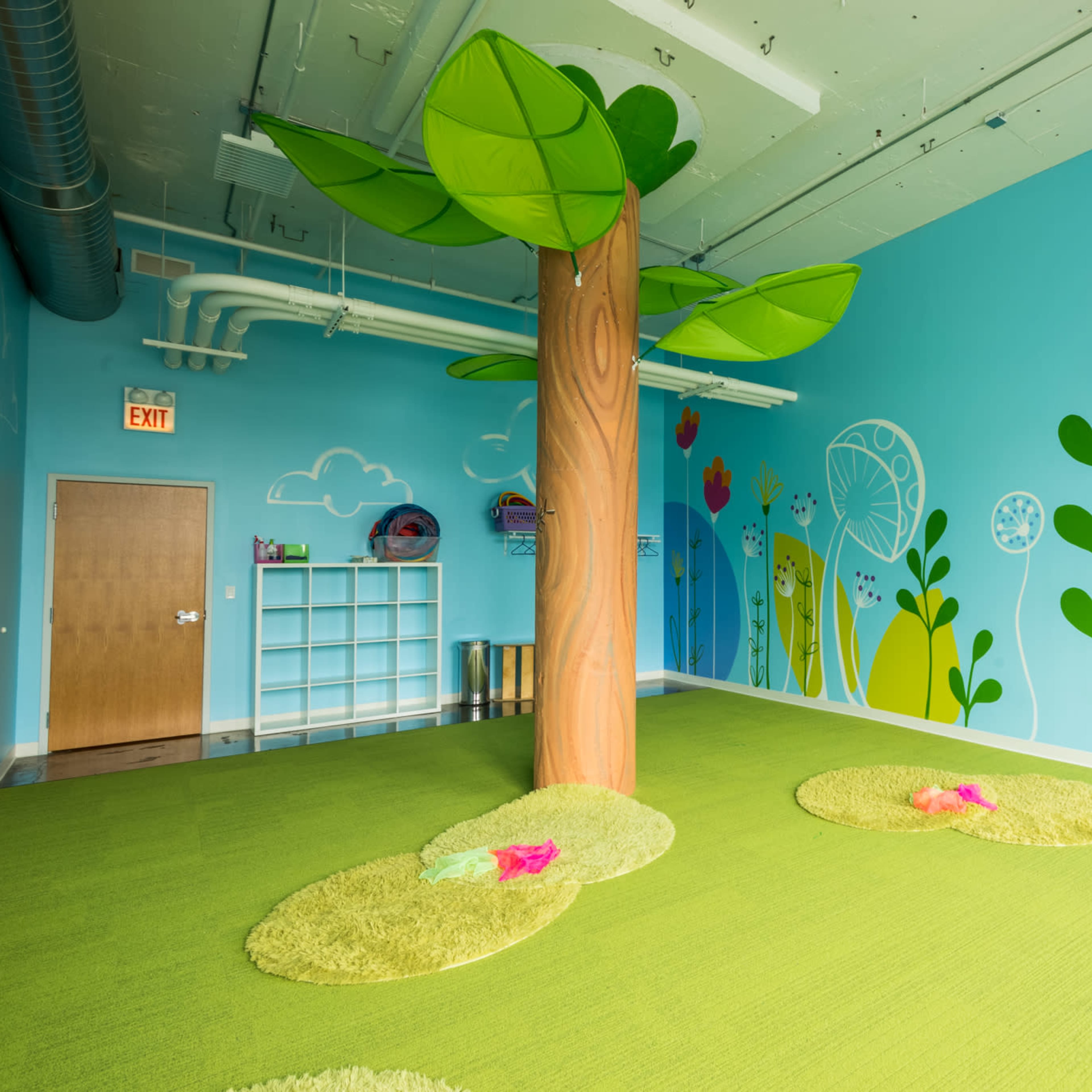 A brightly colored children's playroom with a large tree sculpture, green carpet, and whimsical wall decorations featuring flowers and plants.