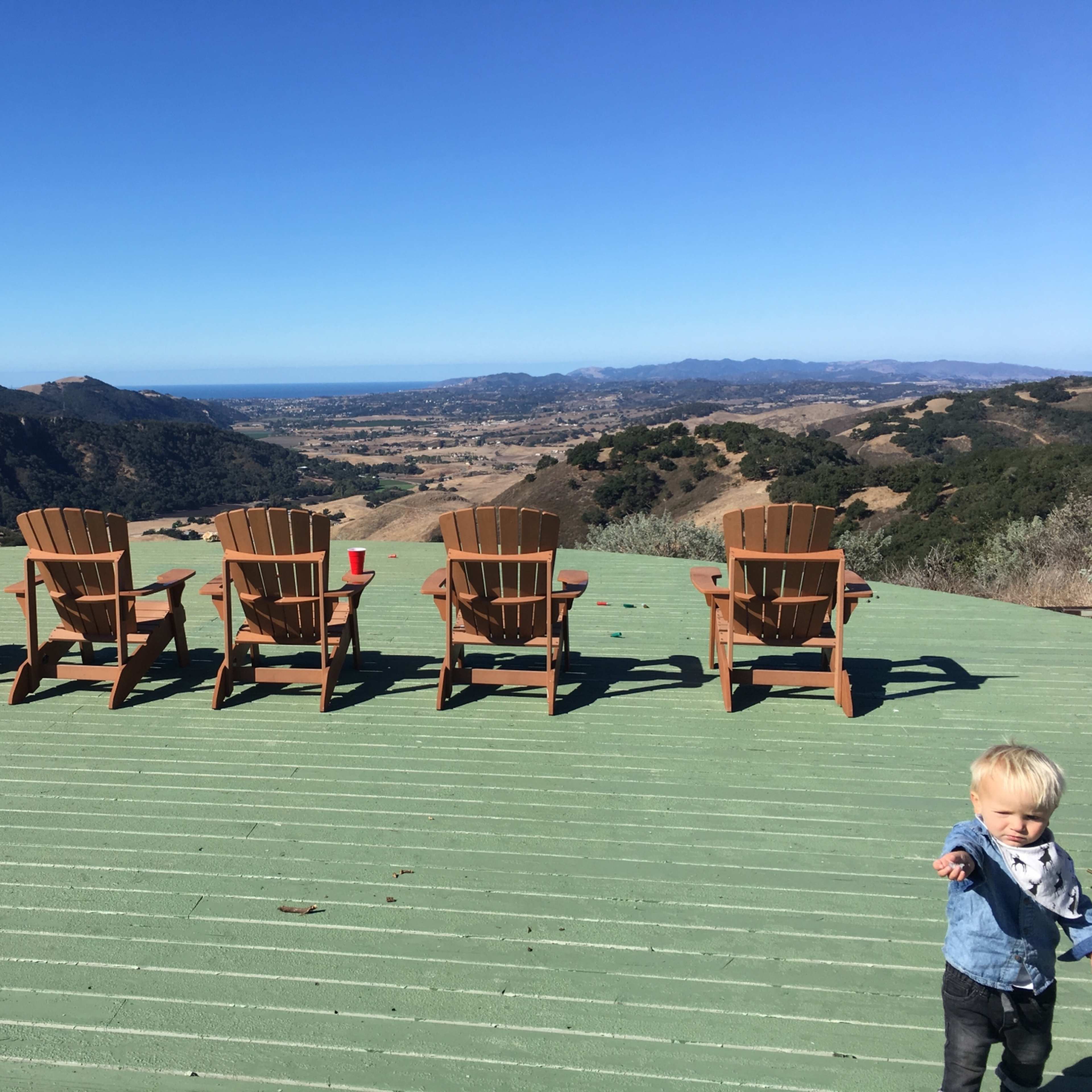 A row of wooden chairs is placed on a green deck overlooking a scenic landscape with hills and distant coastline.