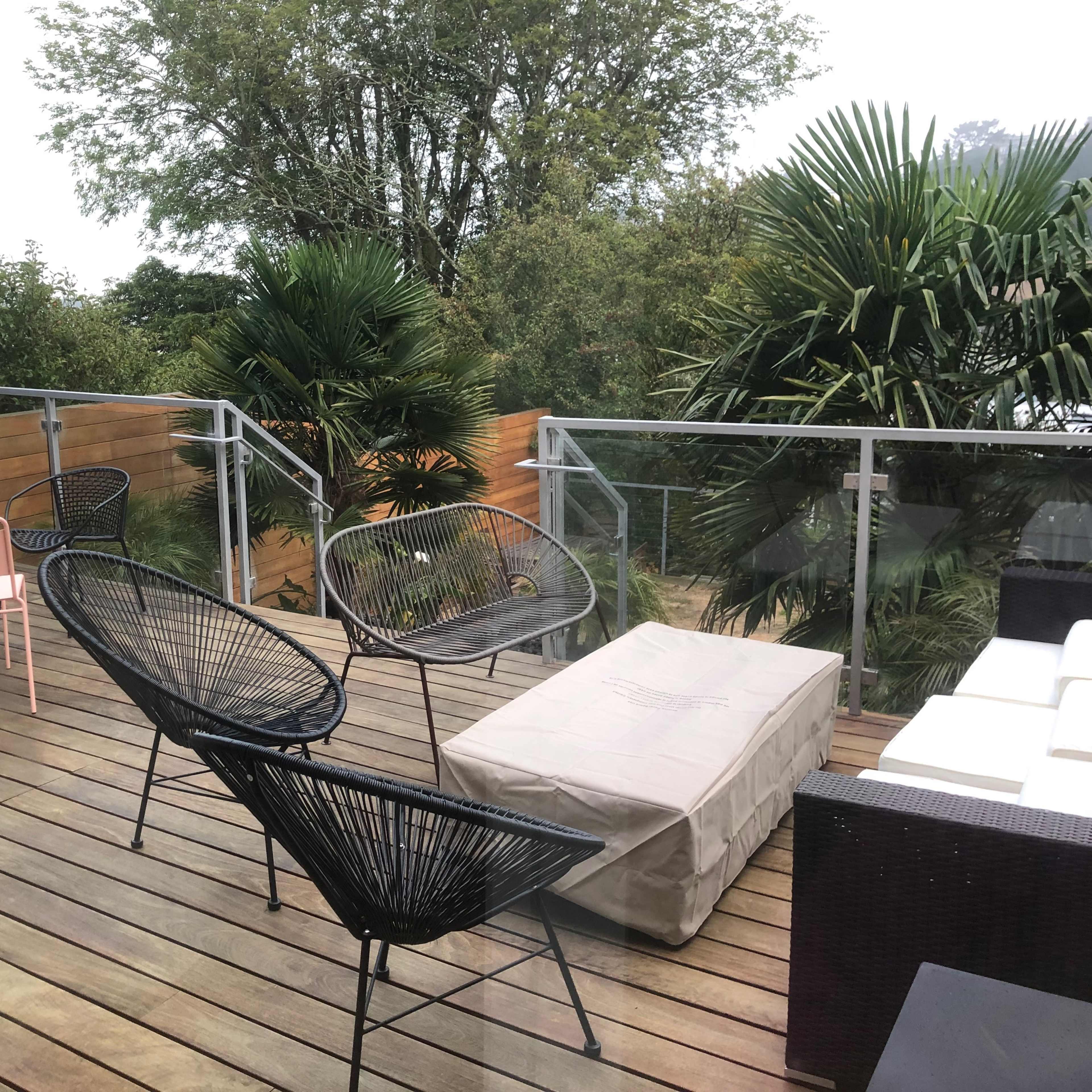 A wooden deck features a variety of outdoor furniture, including black and pink chairs, a coffee table, and tropical plants in the background.