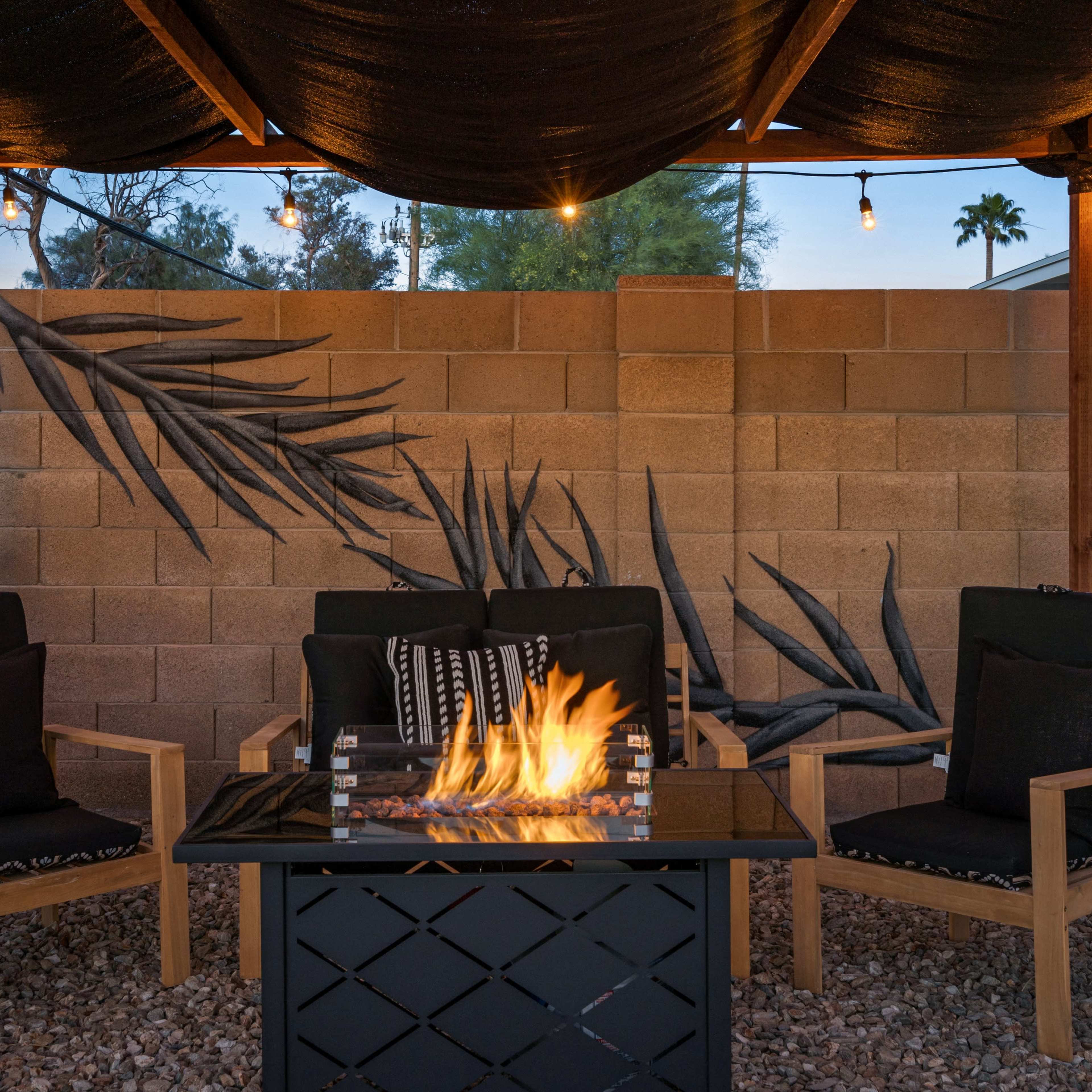 A covered outdoor seating area features two black chairs and a fire table with flames, surrounded by a decorative wall mural and string lights.