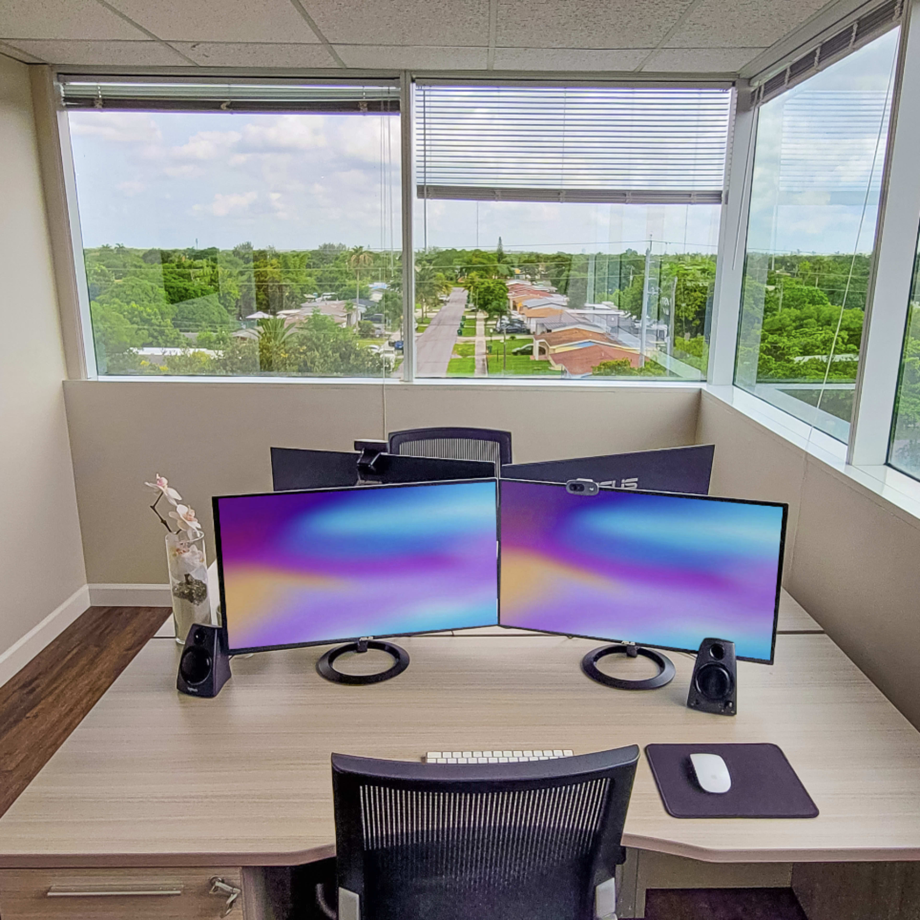 A desk with dual monitors is positioned near a large window overlooking a green landscape and road.