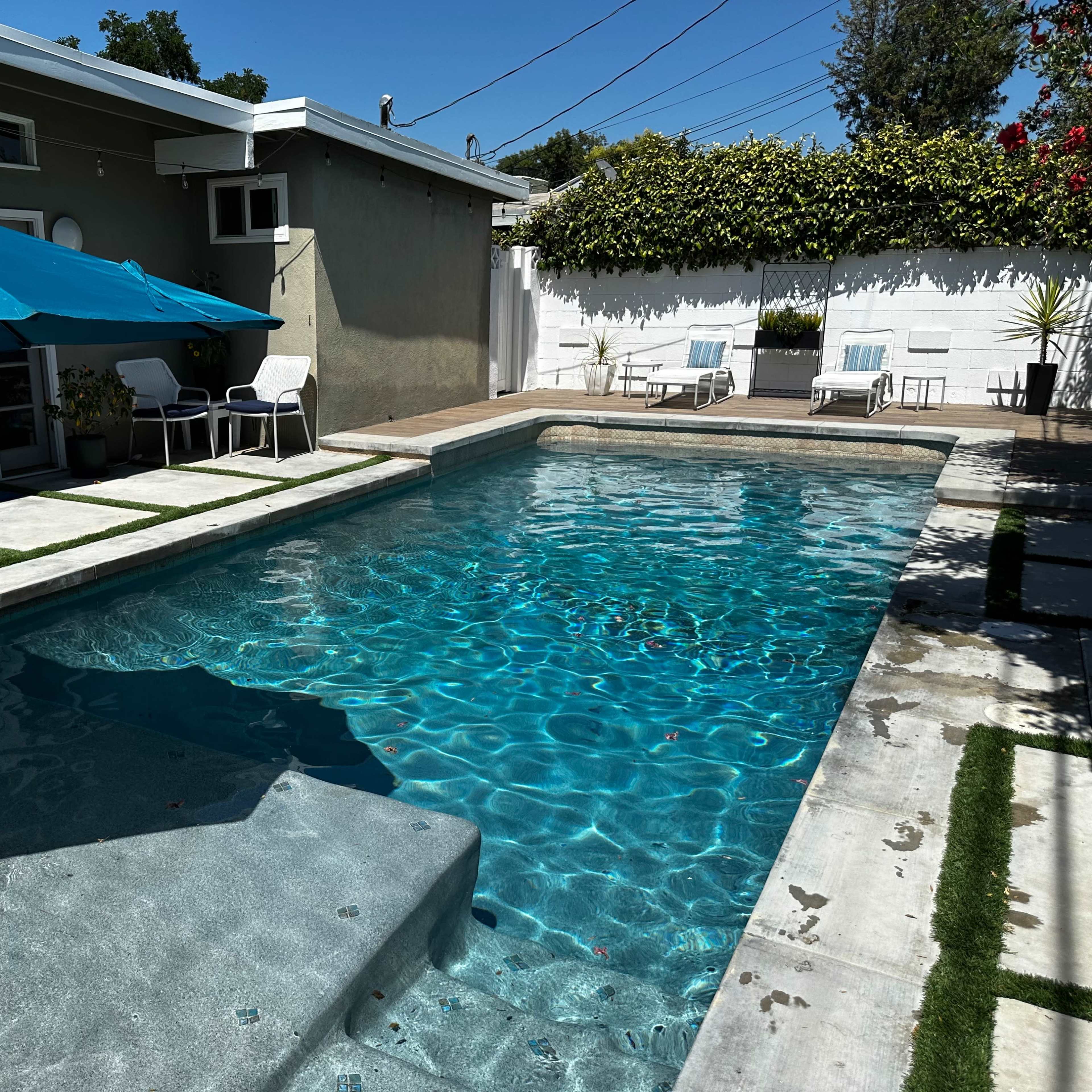 A clear swimming pool surrounded by a patio with lounge chairs and an umbrella.