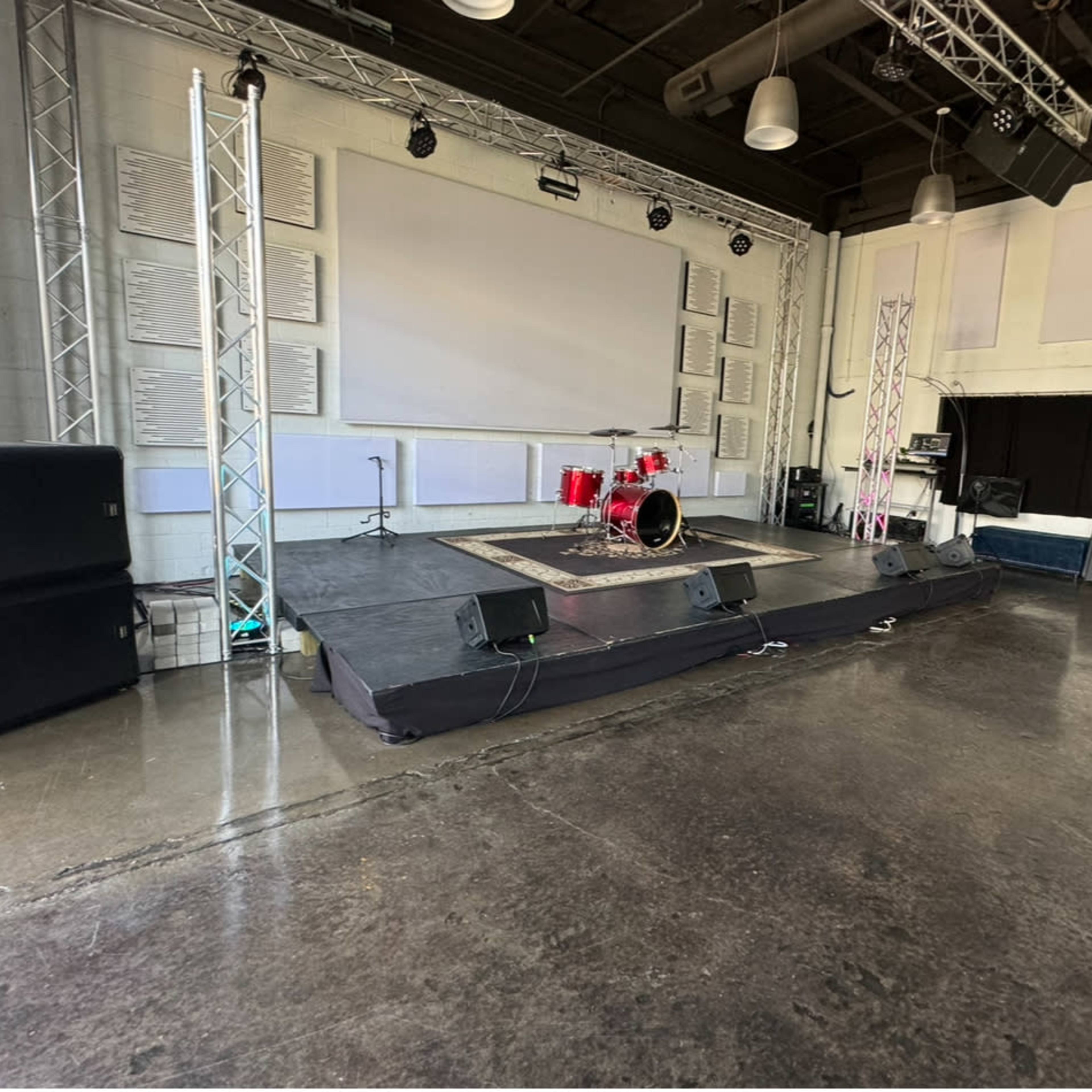 A stage setup in a spacious room, featuring a drum kit and sound equipment against a backdrop of a large screen.