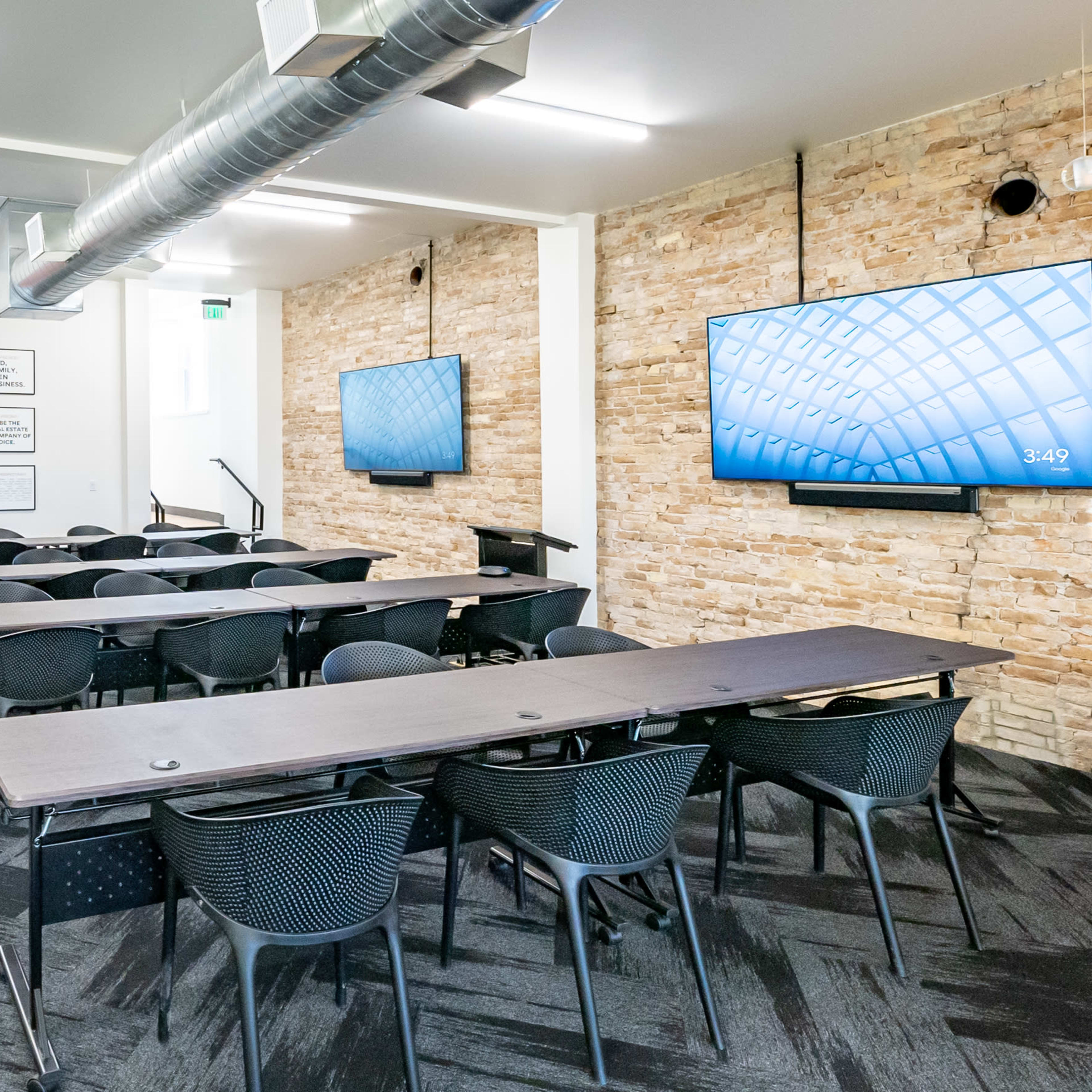A modern classroom with rows of black chairs and tables, equipped with two large screens mounted on the walls and exposed stone accents.