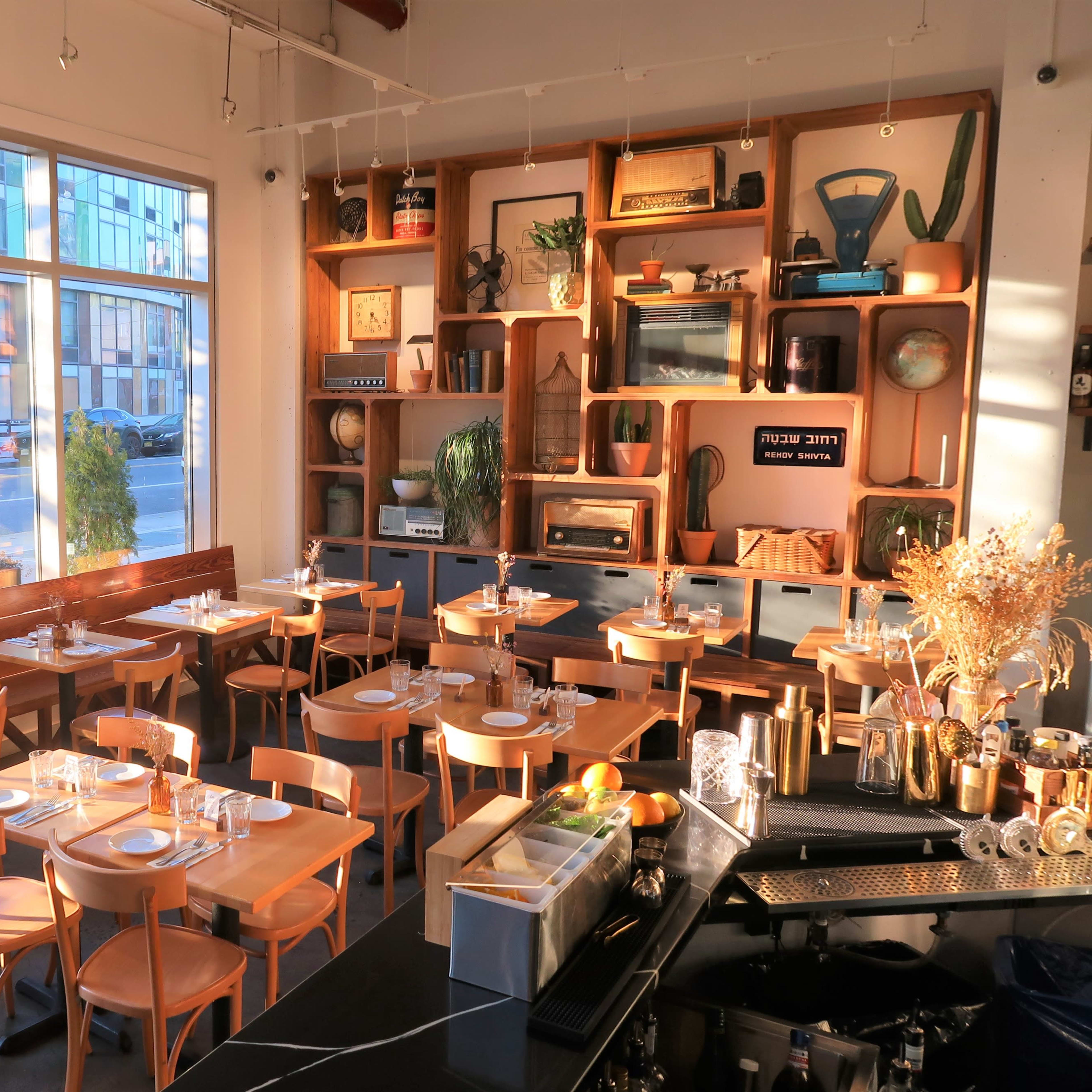 A modern restaurant interior with wooden tables, chairs, and a decorative shelving unit, illuminated by natural light through large windows.