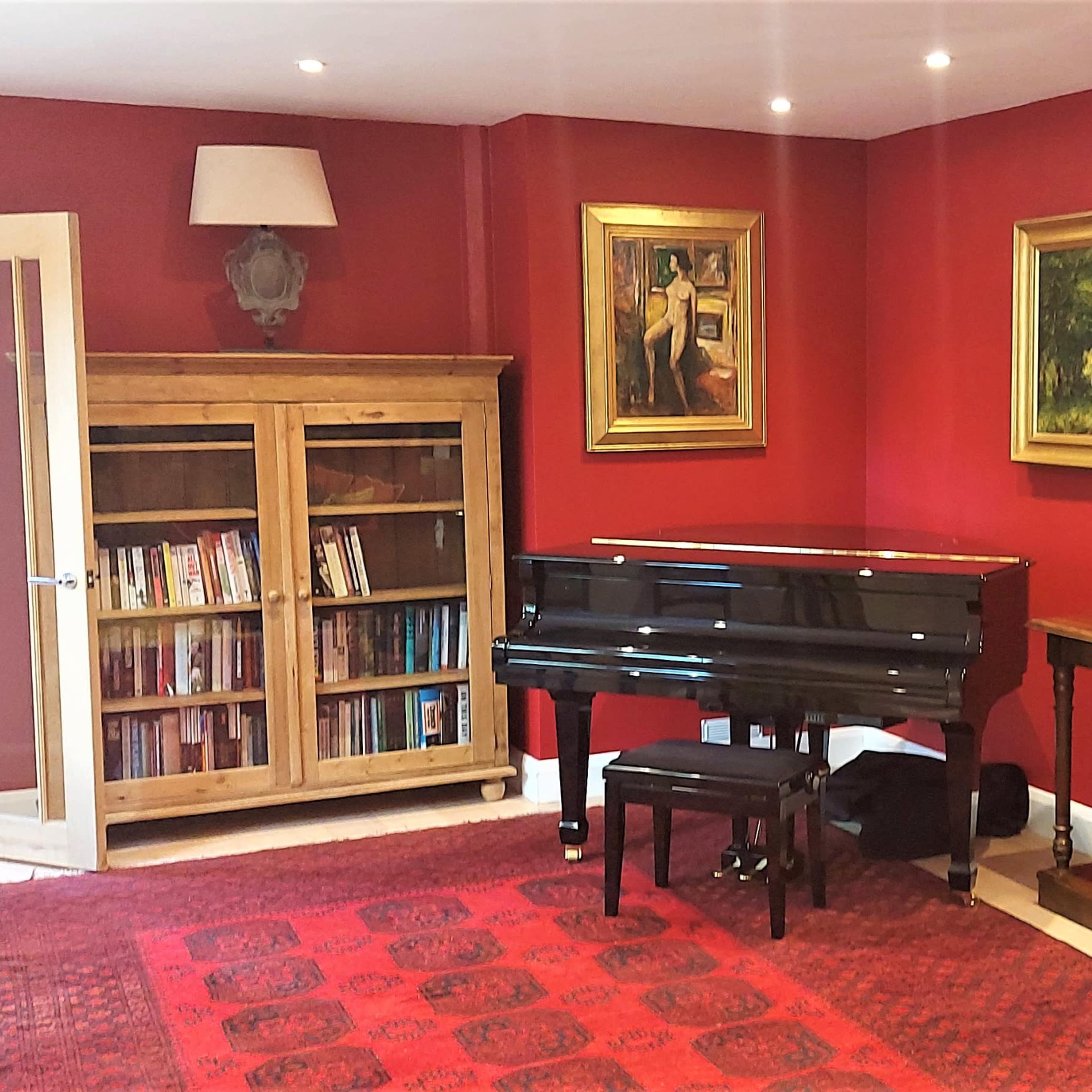 A living room with red walls, featuring a black piano, a wooden bookshelf filled with books, and a side table with a lamp.