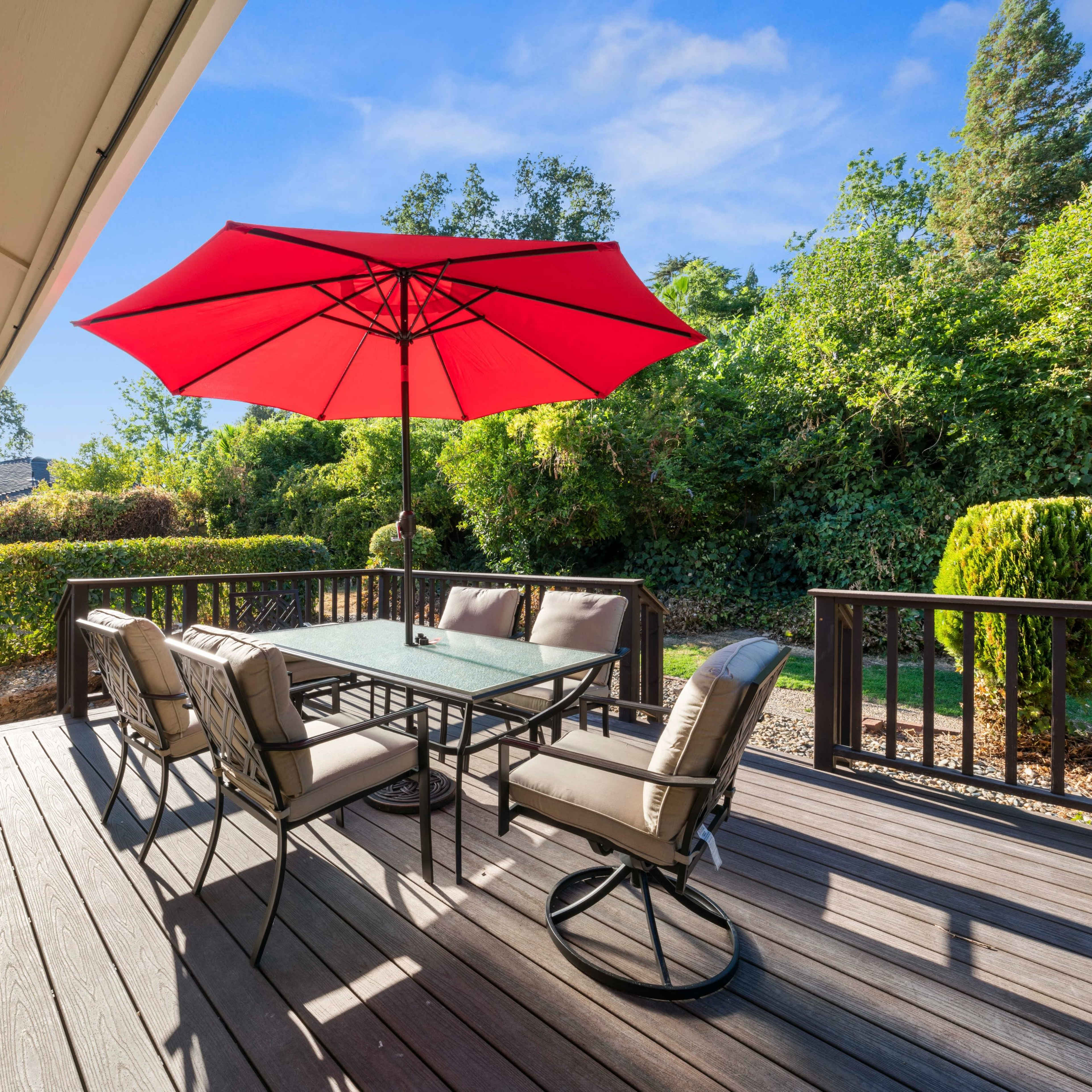 A patio features a table and chairs under a large red umbrella, surrounded by greenery and wooden railings.