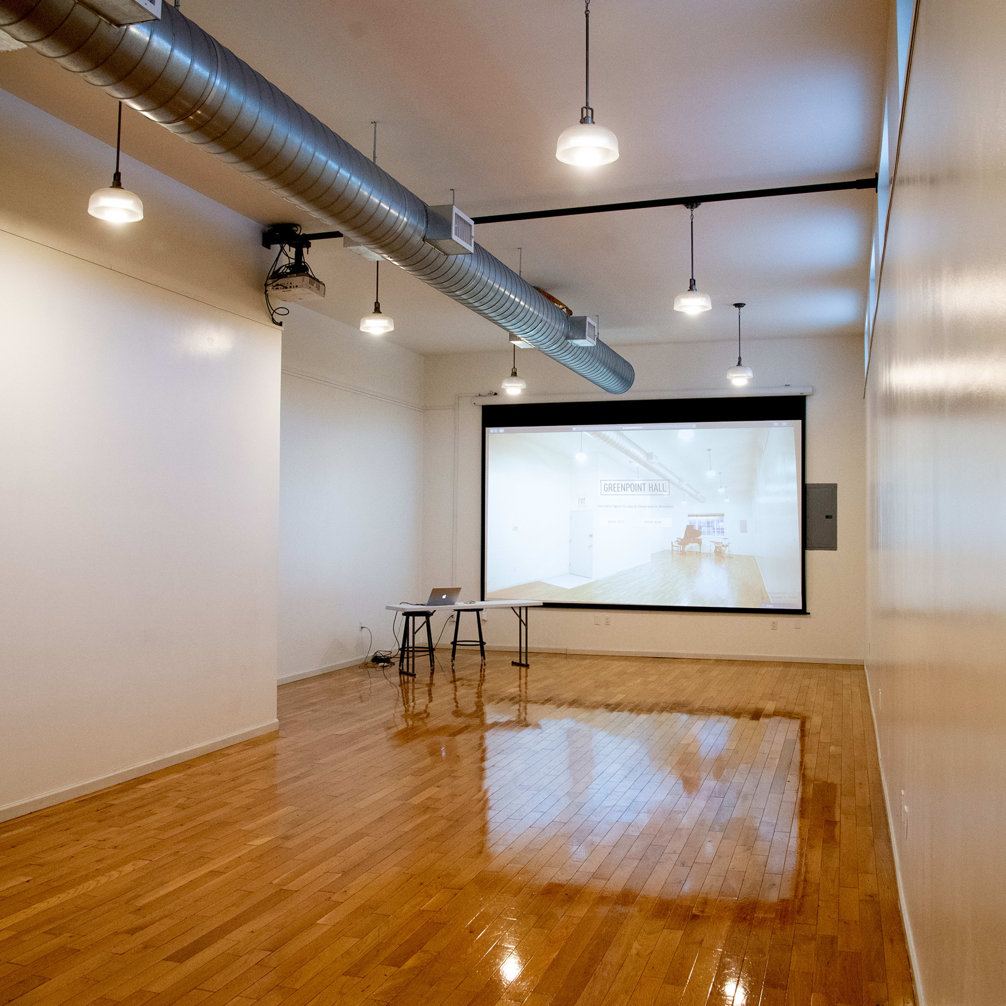 A modern, empty room with wooden floors, a large white screen at the front, and overhead lighting.