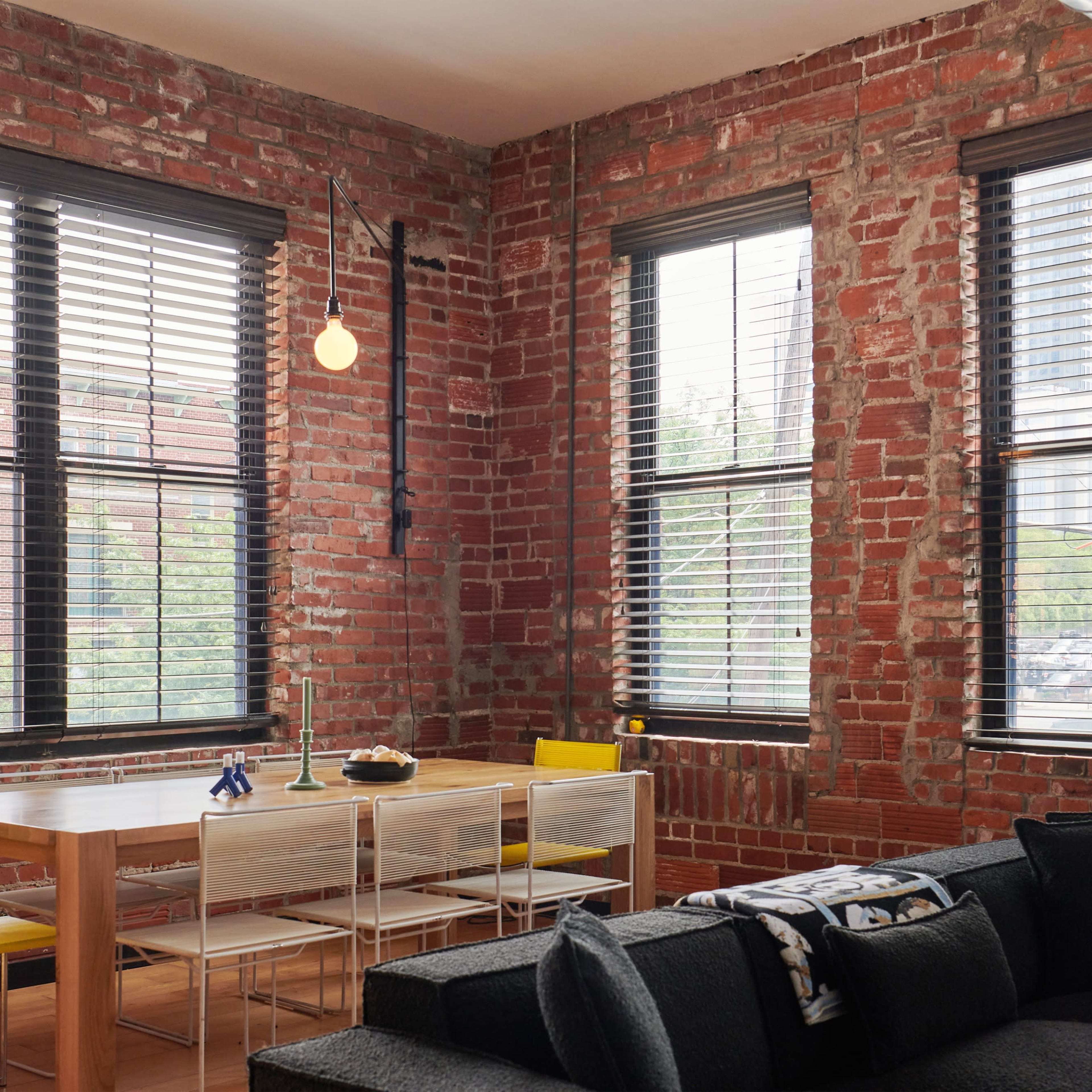 A dining table with yellow and clear chairs is positioned near large windows in a room with exposed brick walls.
