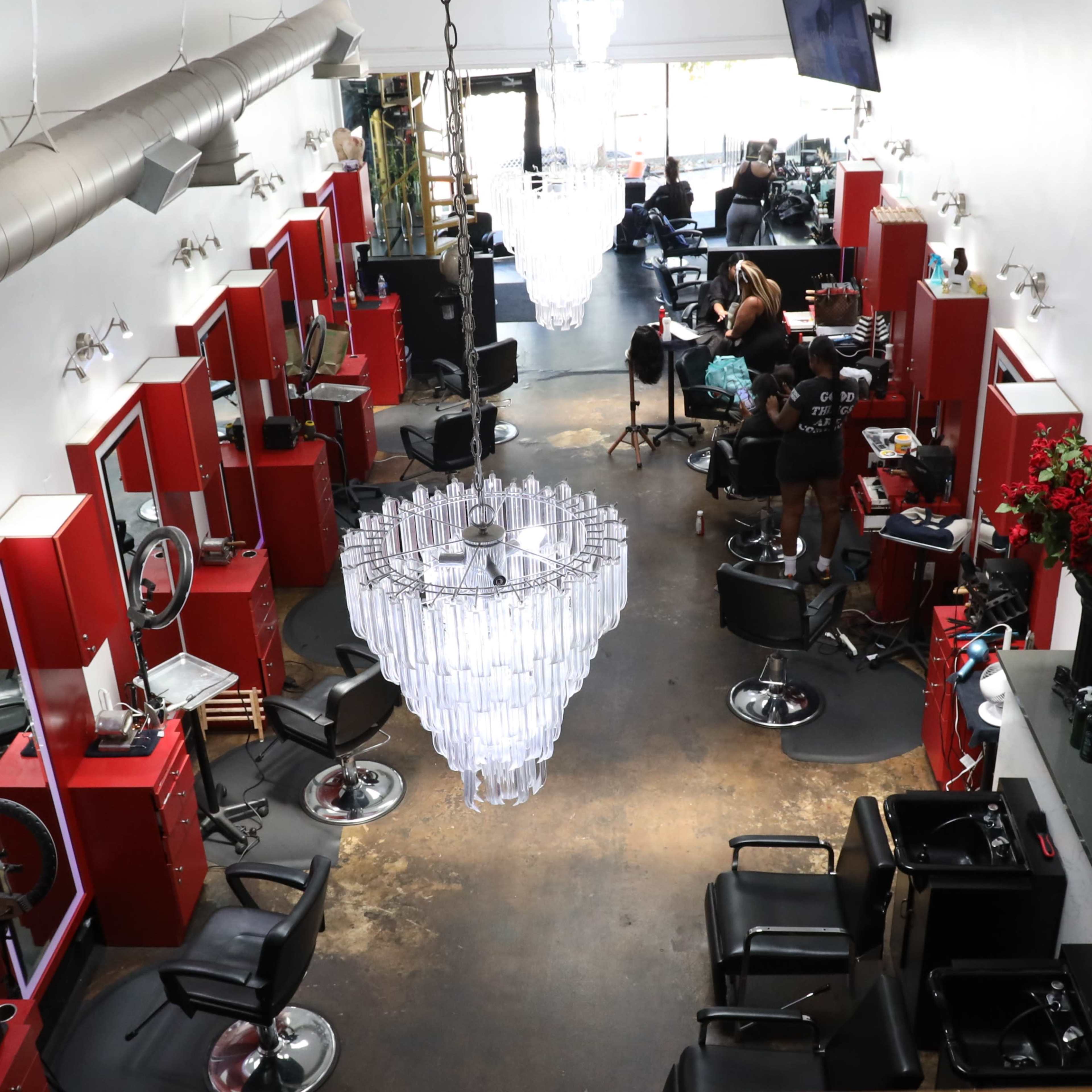 A hair salon with red styling stations, a large chandelier in the center, and stylists working on clients.