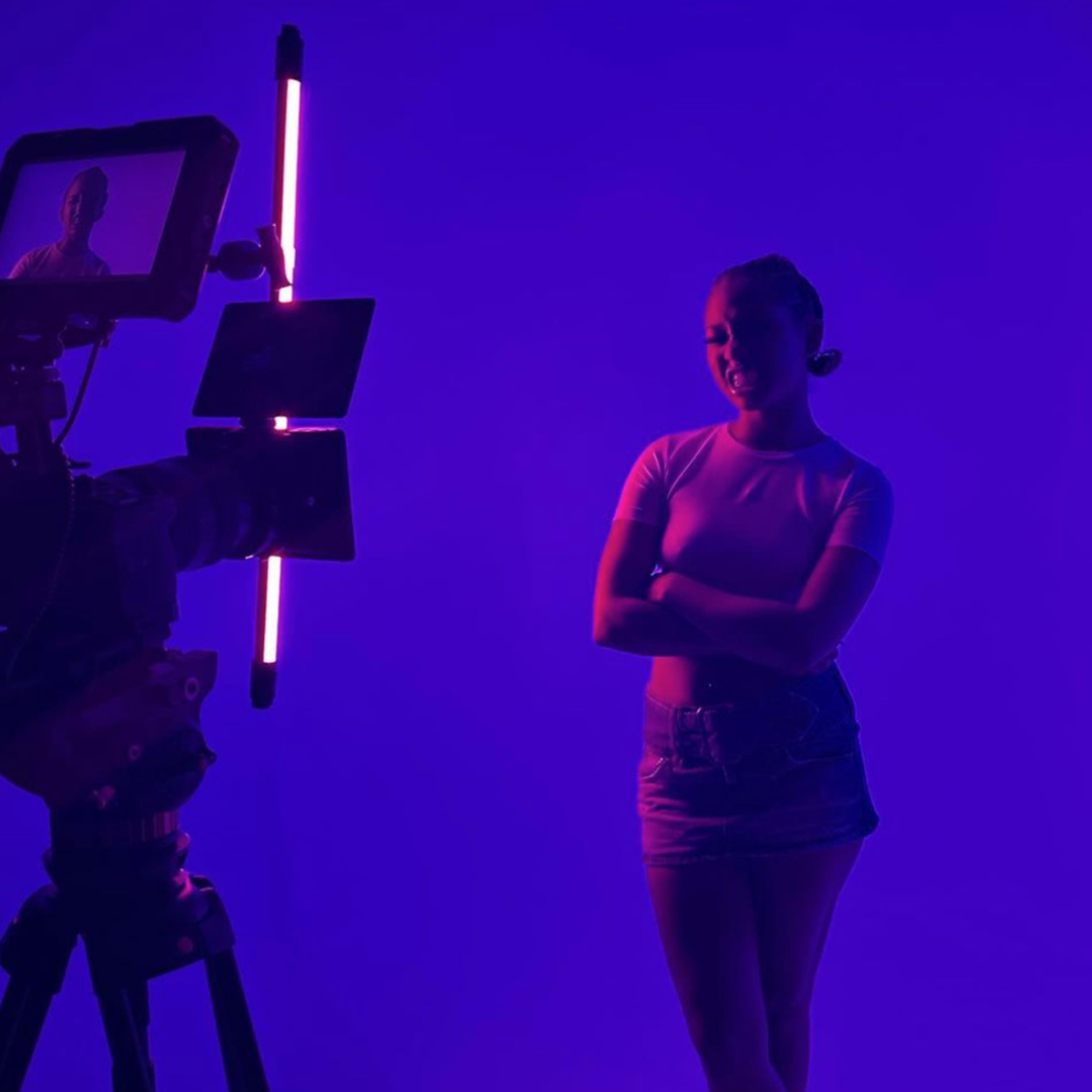 A person stands with arms crossed in front of a camera under purple lighting.