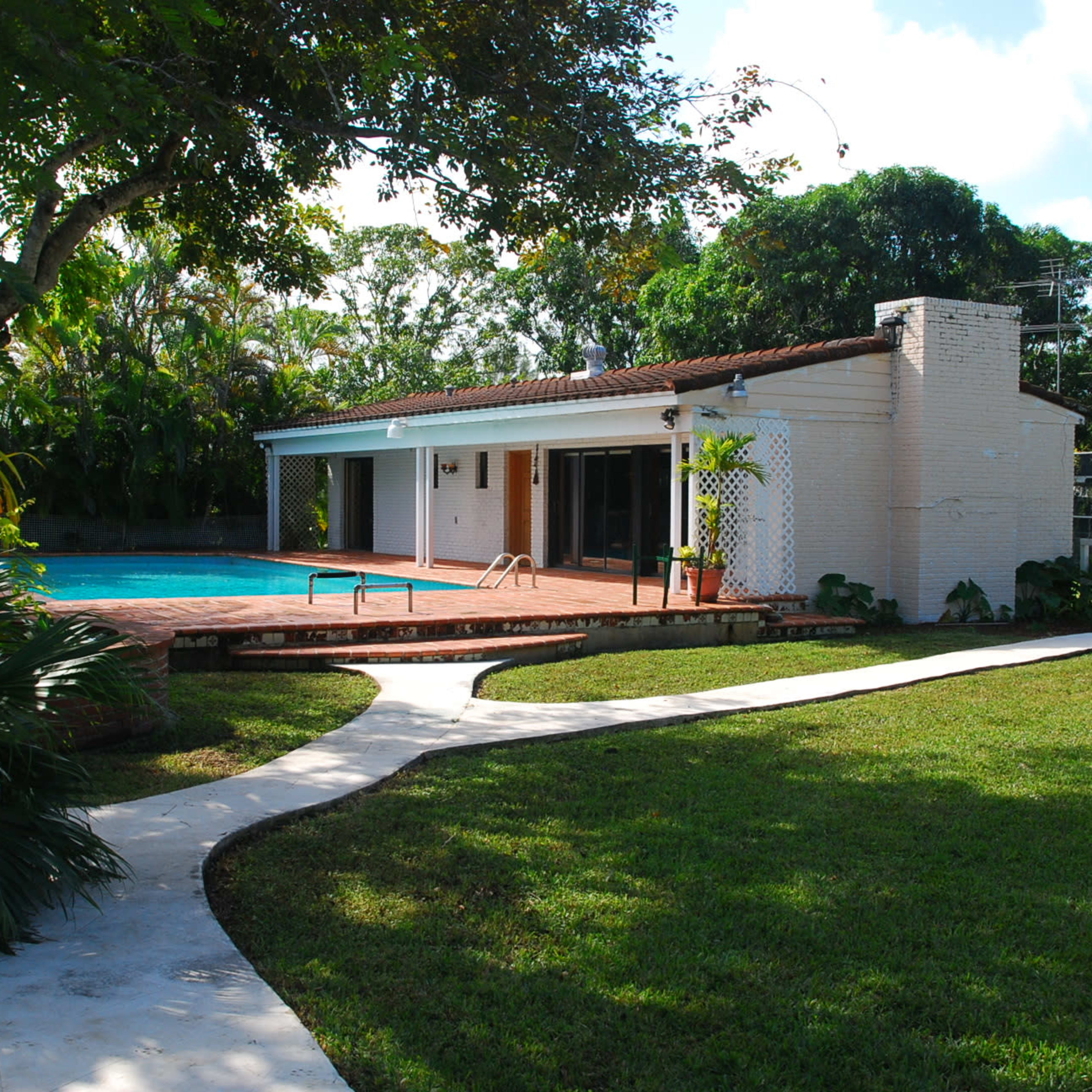 A single-story house with a pool and a paved walkway surrounded by grass and trees.