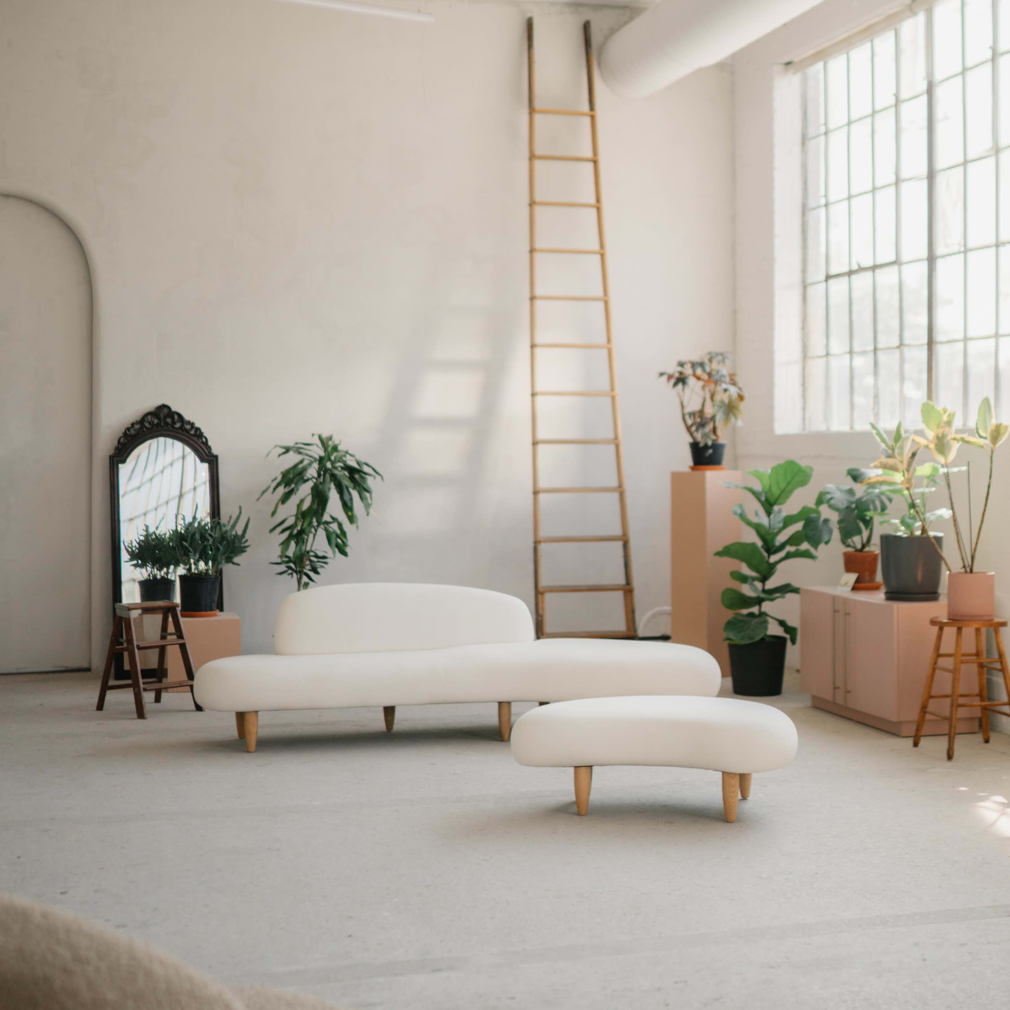 A spacious, light-filled room featuring a white sofa and a small ottoman, with plants and a wooden ladder against a wall near a large window.