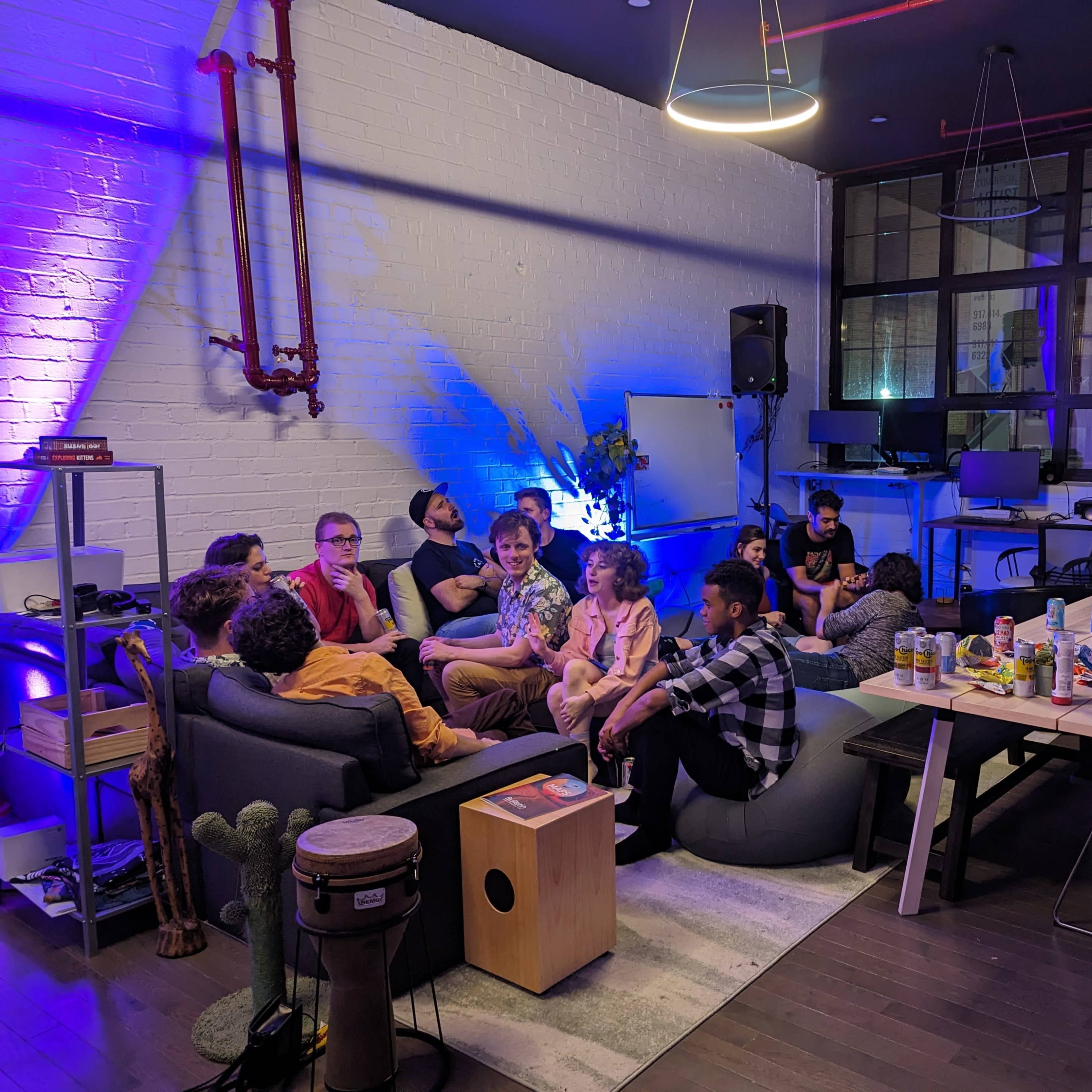 A group of people sits on couches in a well-lit room with modern decor, surrounded by computers and a dining table with snacks.
