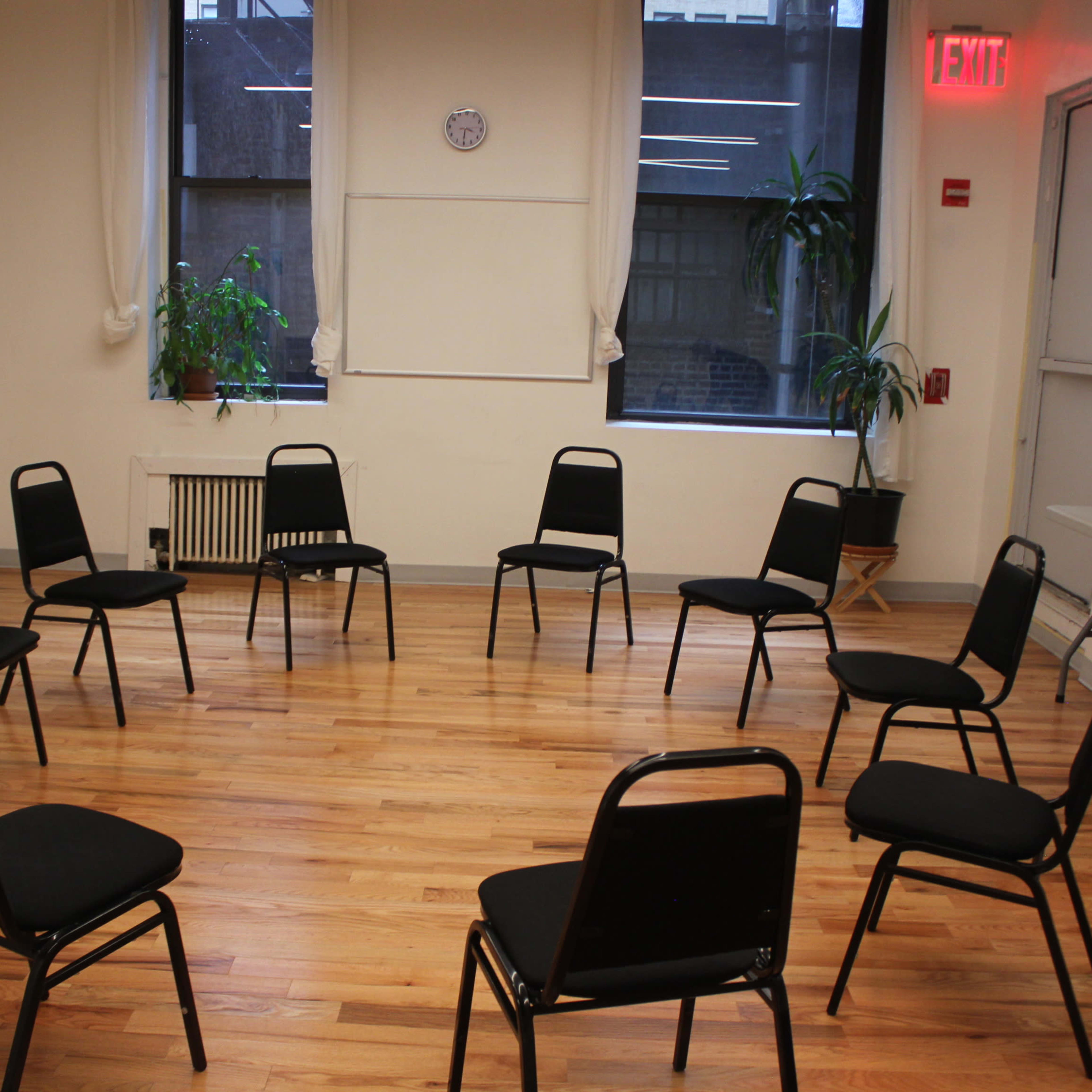 A room with hardwood floors arranged with twelve black chairs in a circle, two windows allowing natural light, and a small table against one wall.