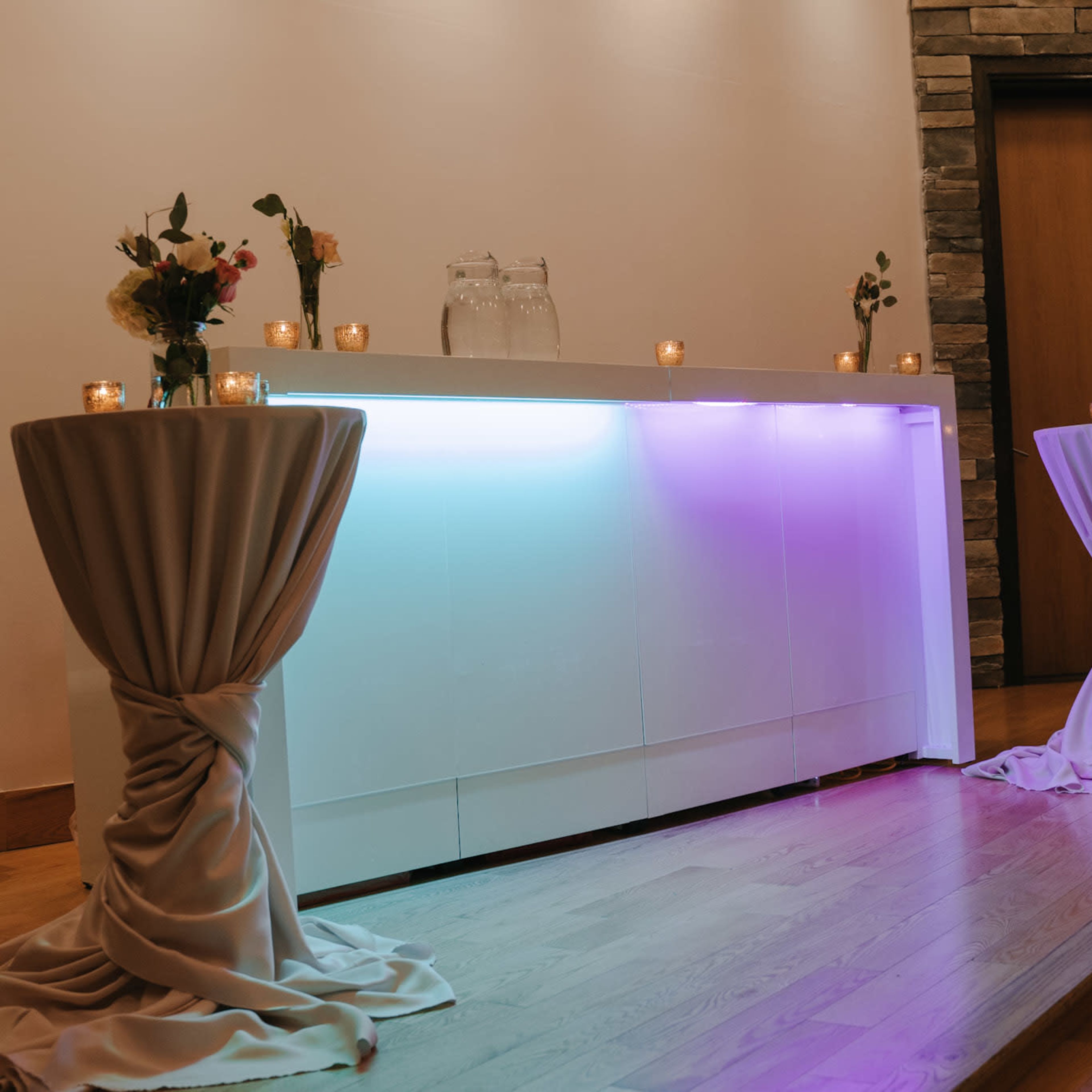 A sleek, illuminated bar is flanked by two draped tables adorned with flowers and candles in a well-lit indoor setting.