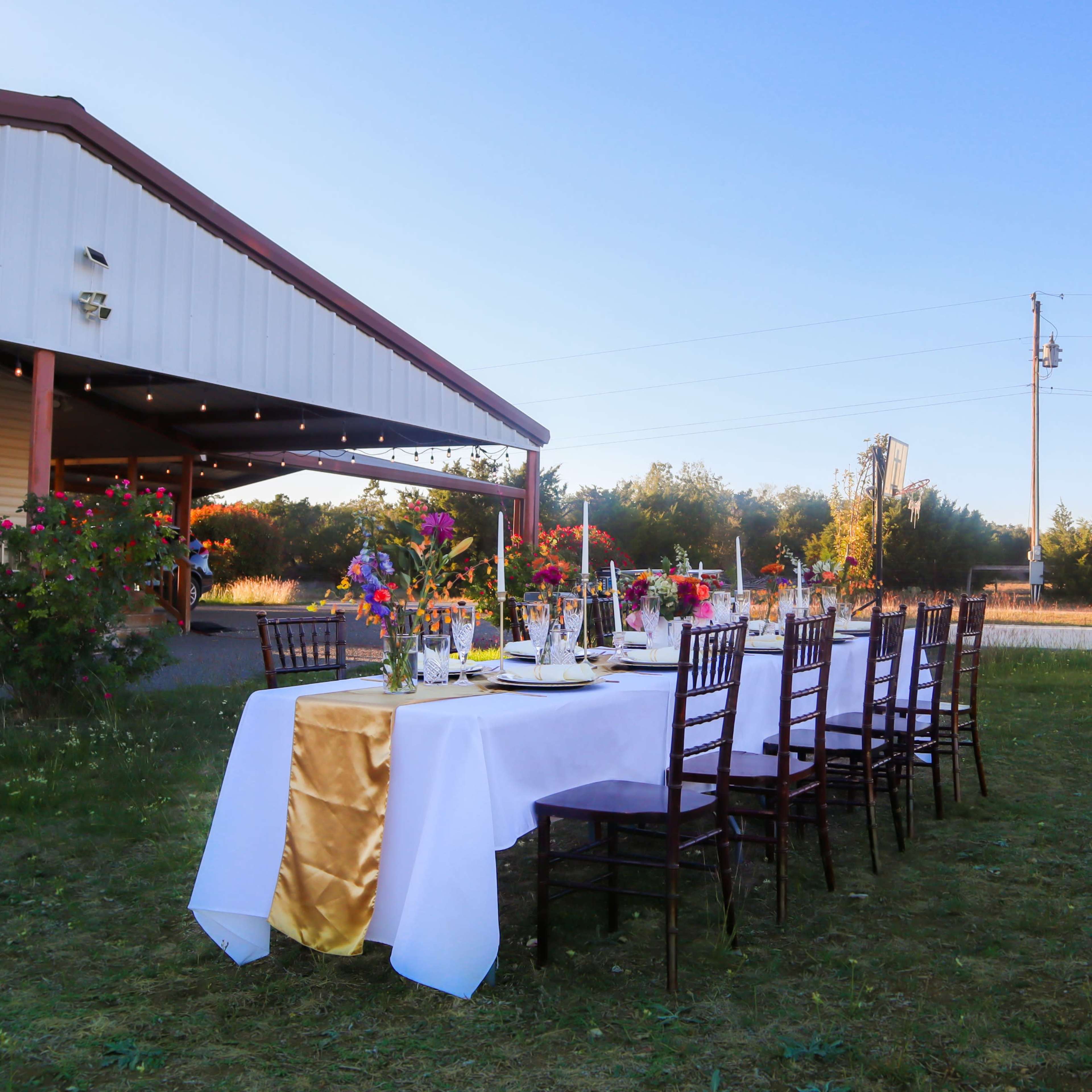 A long dining table is set outdoors, decorated with flowers and tableware, in front of a building and surrounded by open grass and trees.